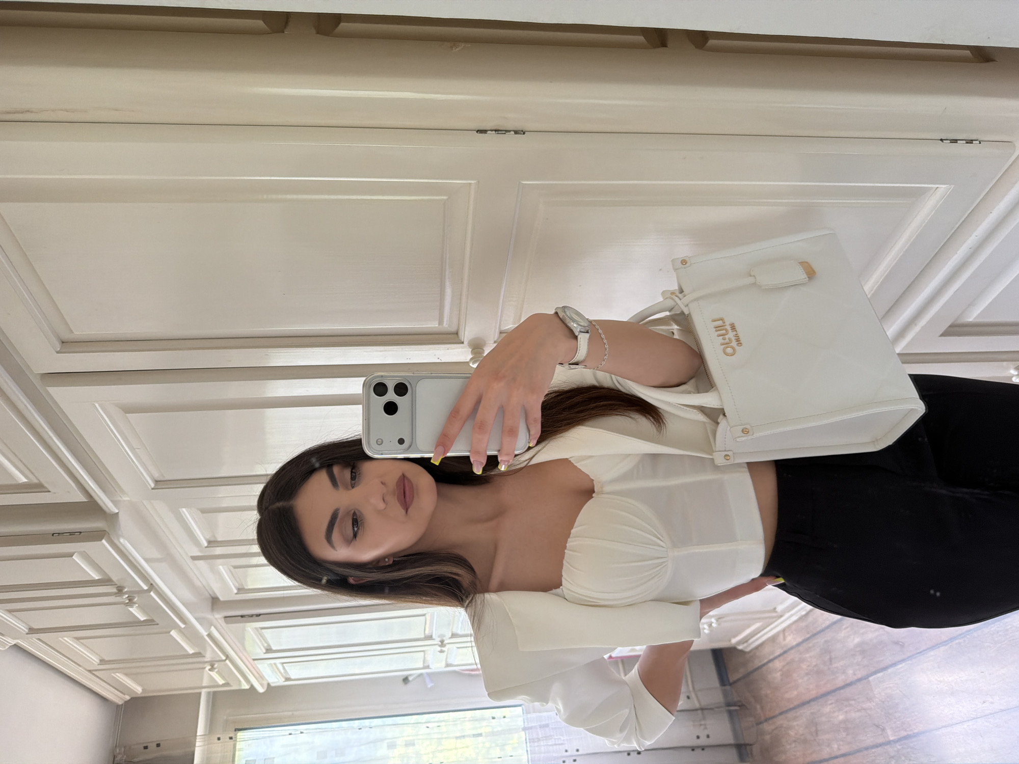 A woman is taking a selfie in a room with white walls and cabinets. She is wearing a white top, black pants, and holding a white handbag. She has brown hair and is wearing a watch and bracelet. The location is not discernible from the image.