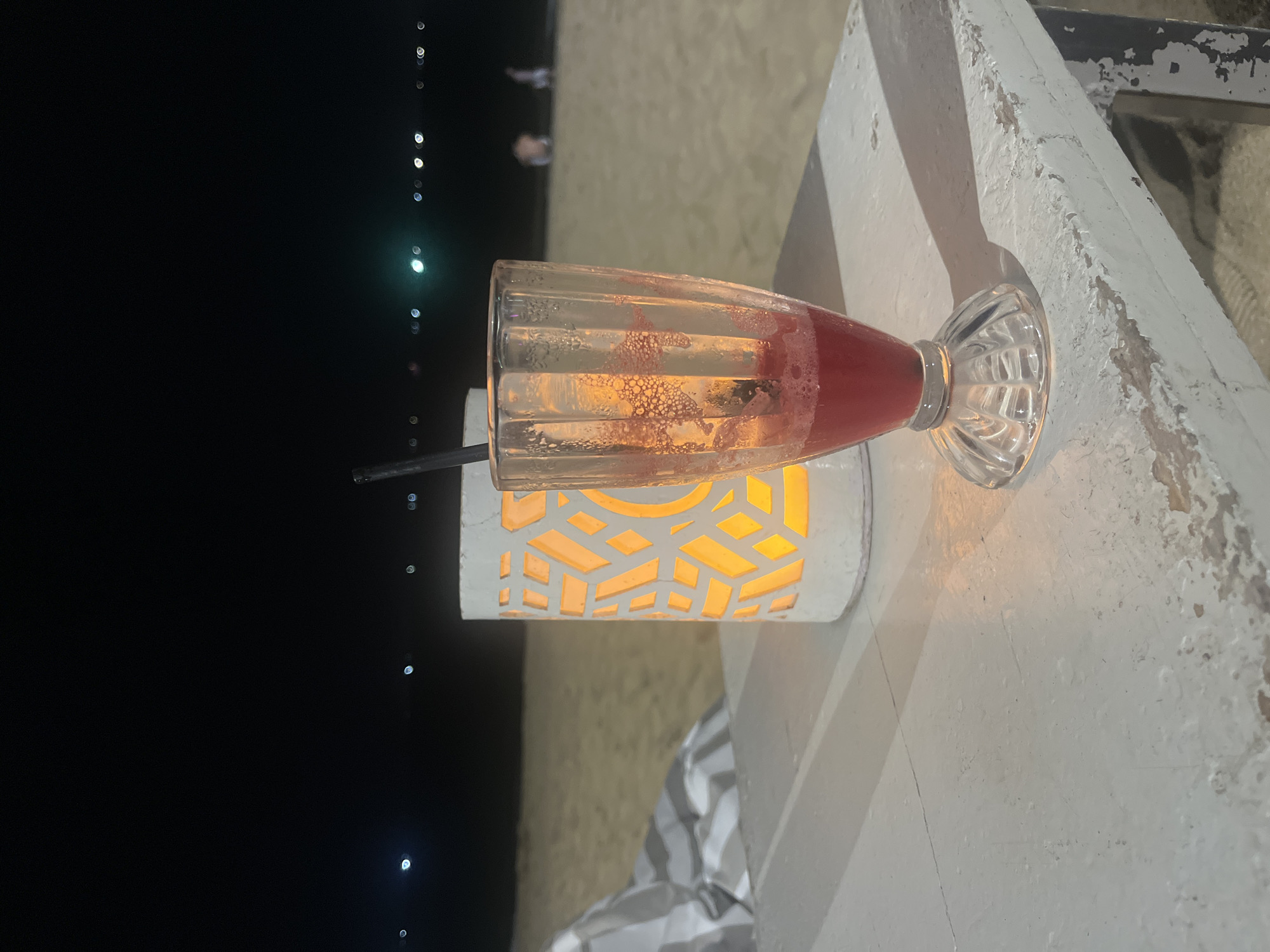 A partially consumed red drink in a stemmed glass sits on a white, weathered table at night. The table is positioned on a sandy beach, with a lit lantern to the left of the glass. The lantern is cylindrical with a cut-out geometric pattern, glowing orange from within. In the background, the dark ocean stretches to the horizon, dotted with distant lights. The sand is visible in the mid-ground. A striped chair is partially visible to the left of the table. The scene suggests a relaxed evening at a beachfront establishment.