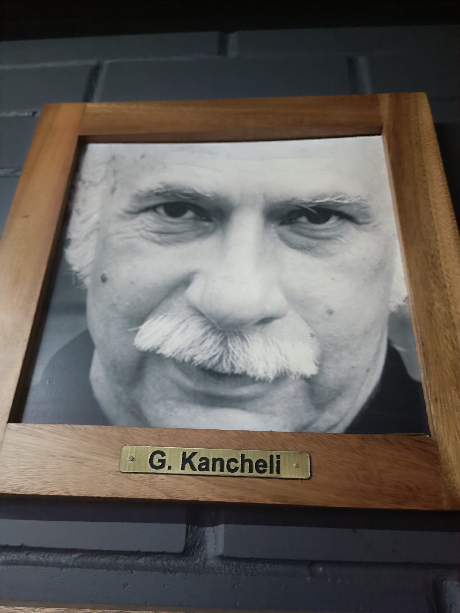 A framed black and white portrait of an older man with a prominent mustache is displayed. The man's face fills most of the frame, and he is looking directly at the viewer. Below the portrait, a brass plaque is affixed to the wooden frame, bearing the name "G. Kancheli." The portrait is hung on a dark, textured wall that appears to be made of bricks.