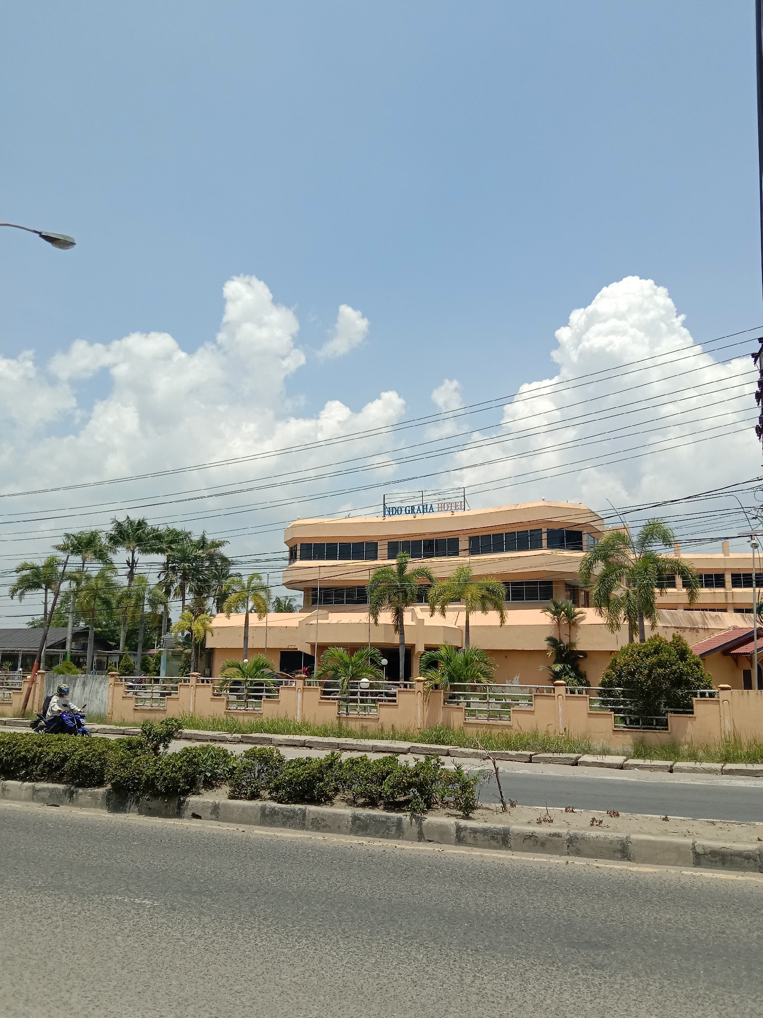 The image depicts a street scene in Lhokseumawe, Indonesia, under a bright, clear blue sky dotted with fluffy white clouds. In the center, a prominent multi-story building, likely a hotel, stands with light peach or beige colored facades and numerous windows. The building features a distinctive curved architecture. On its roof, the blue signage "INDO GRAHA Hotel" is clearly visible.
The hotel is surrounded by lush greenery, including many tall palm trees and smaller bushes. In the foreground, an asphalt road stretches horizontally, divided by a median strip adorned with low green shrubs. A person wearing a helmet is riding a blue motorcycle on the far side of the road, moving towards the left, positioned between the median and the hotel's fence. Overhead, several power lines crisscross the frame, and a street lamp is visible on the far left. The scene suggests a sunny daytime setting in a tropical urban environment.
