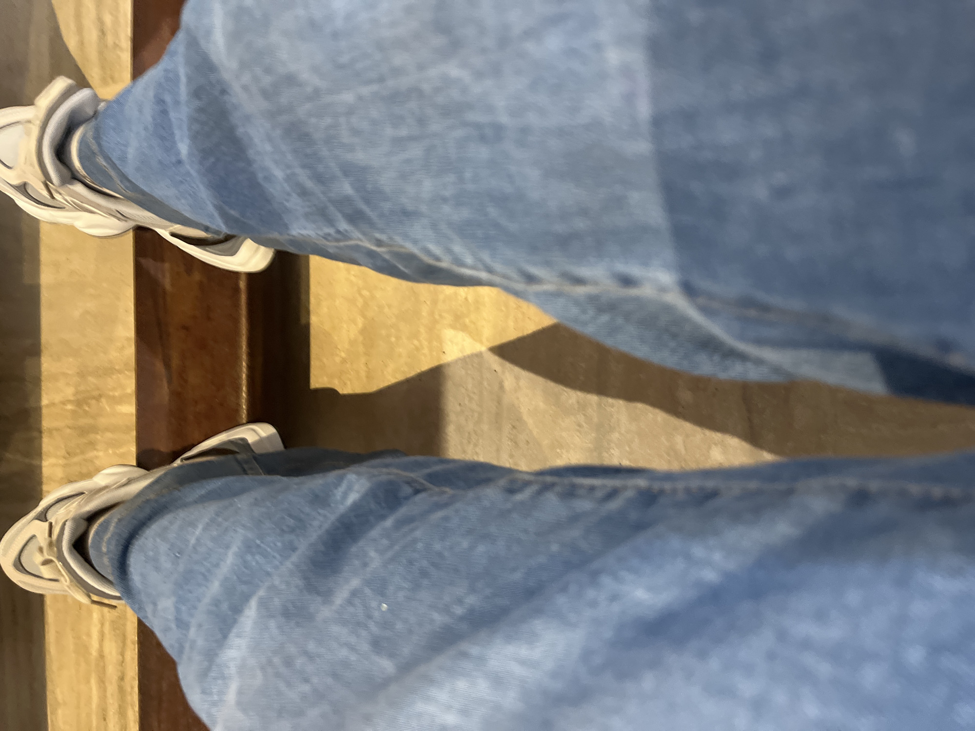 The image displays a first-person, downward perspective of an individual's lower body, specifically their legs and feet, located in Santa Ana, Venezuela. The subject is wearing light blue denim jeans that exhibit characteristic fabric folds and variations in color, indicative of wear. On the feet are light-colored, possibly white or off-white, athletic shoes with visible design elements and robust soles. The feet are positioned on a light tan or beige floor surface, which appears mottled, suggesting materials such as concrete or tile. In the immediate foreground, a two-tiered horizontal structure is visible: an upper section of lighter brown, possibly wood or stone, and a lower section of darker brown with a glossy finish, potentially polished wood. Shadows are sharply cast from the legs and shoes onto the floor, indicating a distinct, likely overhead, light source. No other individuals or specific actions are discernible.