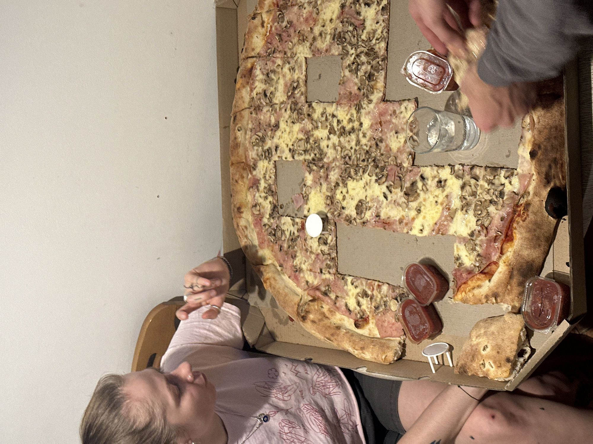 Two individuals are present. One, a young woman, is seated with a pizza box in her lap, while another person is reaching into the box. The pizza is topped with ham and mushrooms, and there are several small containers of what appears to be sauce and a small table in the box. A glass of liquid is also in the box. The background consists of a plain, off-white wall. This image was taken in Novi Sad, Serbia.