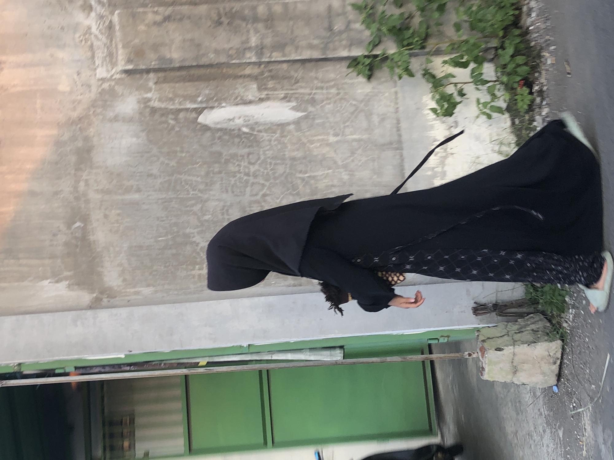 In Glagah Arum, Indonesia, a person wearing a black abaya and hijab walks on a paved surface next to a rough concrete wall. They are carrying a small object in their right hand. To the left of the person, a green metal door is visible, and to the right, a small bush with green leaves grows near the wall. The ground appears to be asphalt or concrete, with some debris and a concrete structure. The lighting suggests it is daytime, with some shadows present.