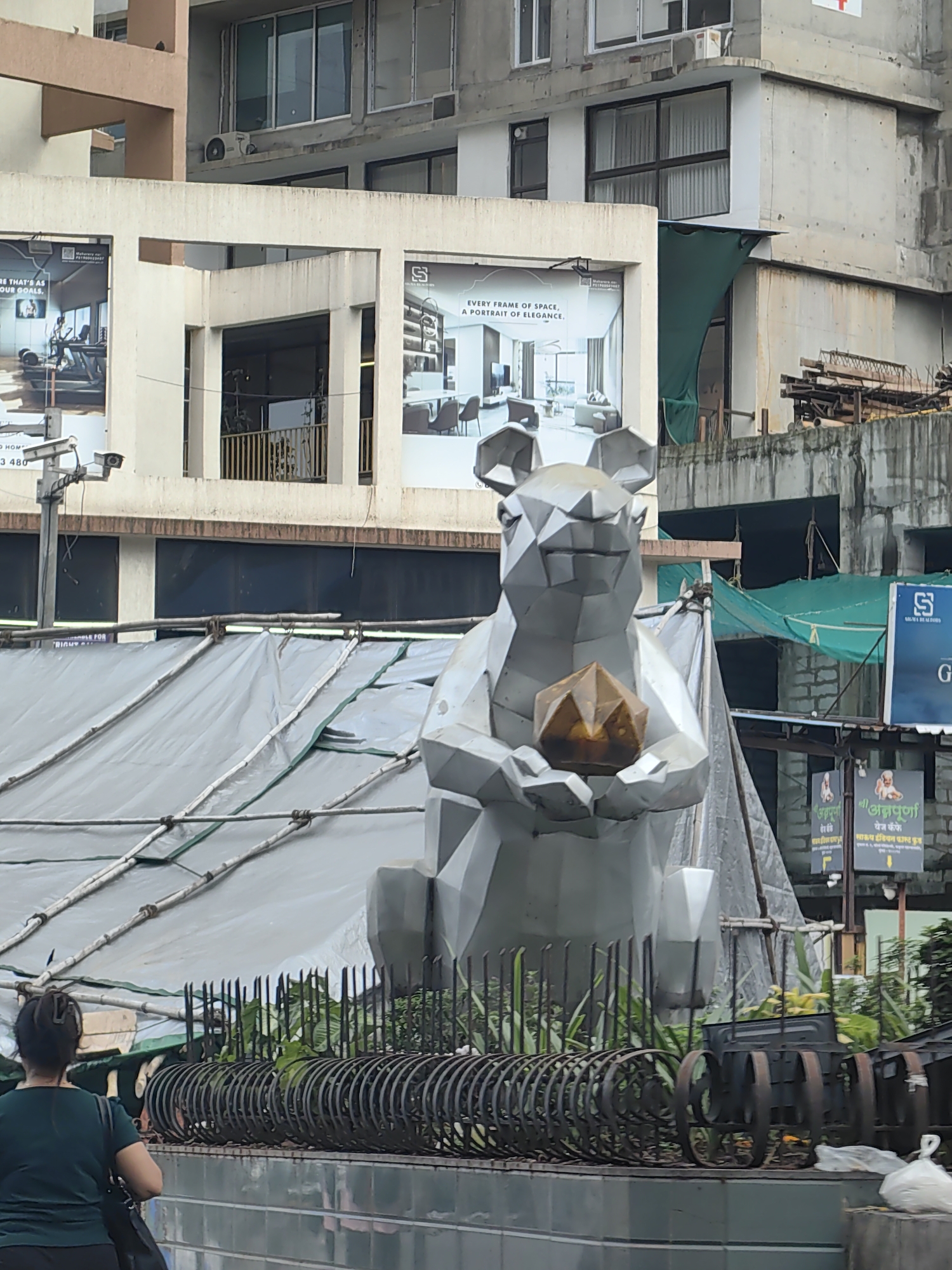 The image depicts a street scene in Mumbai, India. In the foreground, a woman wearing a green t-shirt and dark pants is visible from the back, carrying a dark bag. She appears to be walking. In the middle ground, a large, geometric metal sculpture of a rat holding a faceted amber-colored object is the focal point. Behind the sculpture, a large area is covered with a gray tarp supported by bamboo poles, suggesting ongoing construction or protection of an area. Further back, buildings rise, some appearing to be under construction with exposed concrete and scaffolding. Billboards are visible on the buildings, one advertising what appears to be a gym and another for a real estate company. The overall environment suggests an urban setting with commercial and residential elements, and evidence of development.