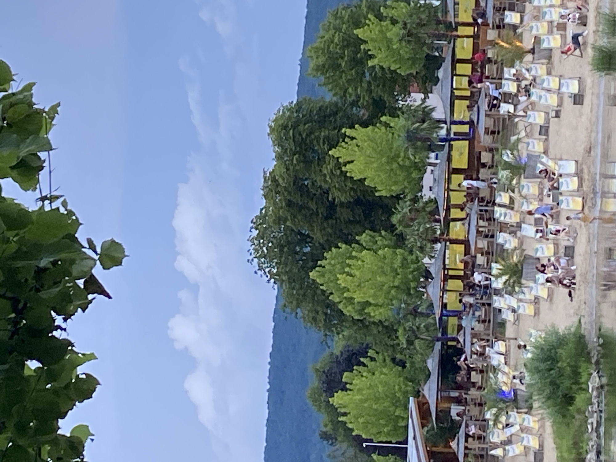 This is a photograph taken at the Weserufer in Rinteln, Germany, specifically at a beach bar. The image captures a lively outdoor scene with numerous people relaxing on lounge chairs on a sandy beach. The beach is equipped with umbrellas providing shade for the seating areas. In the background, there are several structures with thatched roofs and open-sided seating areas, suggesting a restaurant or bar establishment. Green trees are visible behind these structures, and a distant, tree-covered hill forms the backdrop under a cloudy sky. The foreground shows the edge of the Weser river.