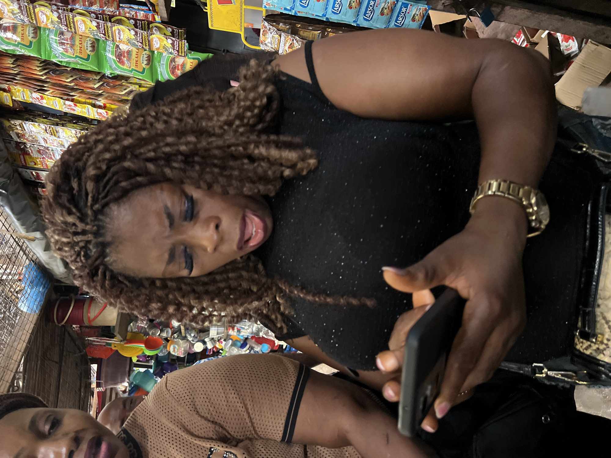 A medium shot shows three people in a market in Nkpor, Nigeria. A woman in the foreground, wearing a black, sparkly sleeveless top and a gold watch, is holding a smartphone and looking at it with her mouth open in surprise. To her left, a woman in a brown, mesh-like shirt is partially visible, looking towards the left. Behind them, a man's head is visible between the two women. The background is filled with shelves of packaged goods, colorful buckets, and other market items, suggesting a bustling retail environment.