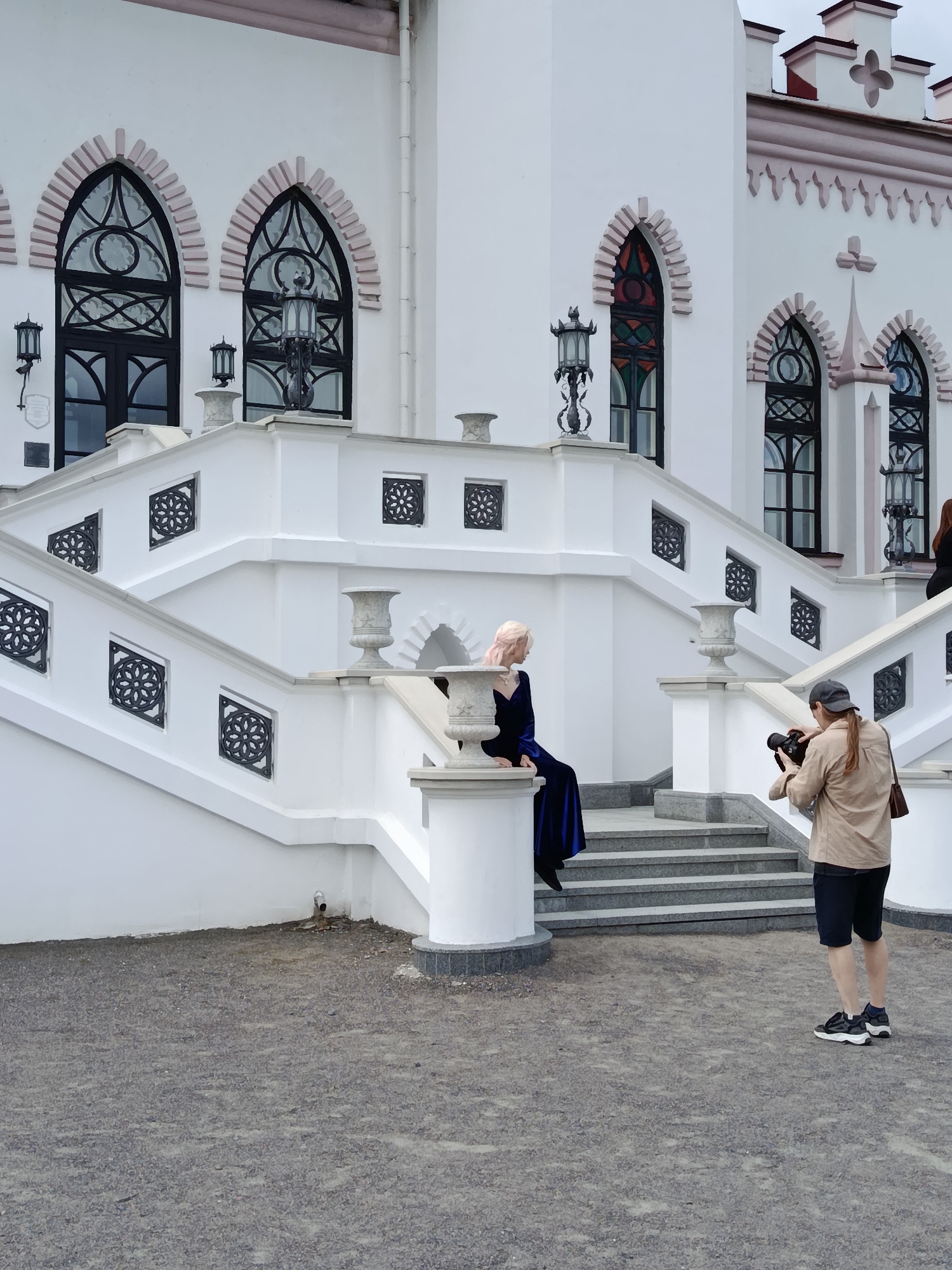 A woman with blonde hair and pink highlights, wearing a long, dark blue velvet dress, is seated on the edge of a white stone structure on a wide, multi-tiered staircase. She is looking to her left. A person wearing a tan jacket, black shorts, a baseball cap, and sneakers is standing on the gravel ground at the bottom of the stairs, holding a camera and appearing to take a photo of the seated woman. The staircase and the building behind it are painted white with decorative pink trim around arched windows. The windows feature dark frames and intricate metalwork. The ground in front of the stairs is covered in small grey gravel. The scene appears to be in front of the Kosava Castle in Kosava, Belarus.