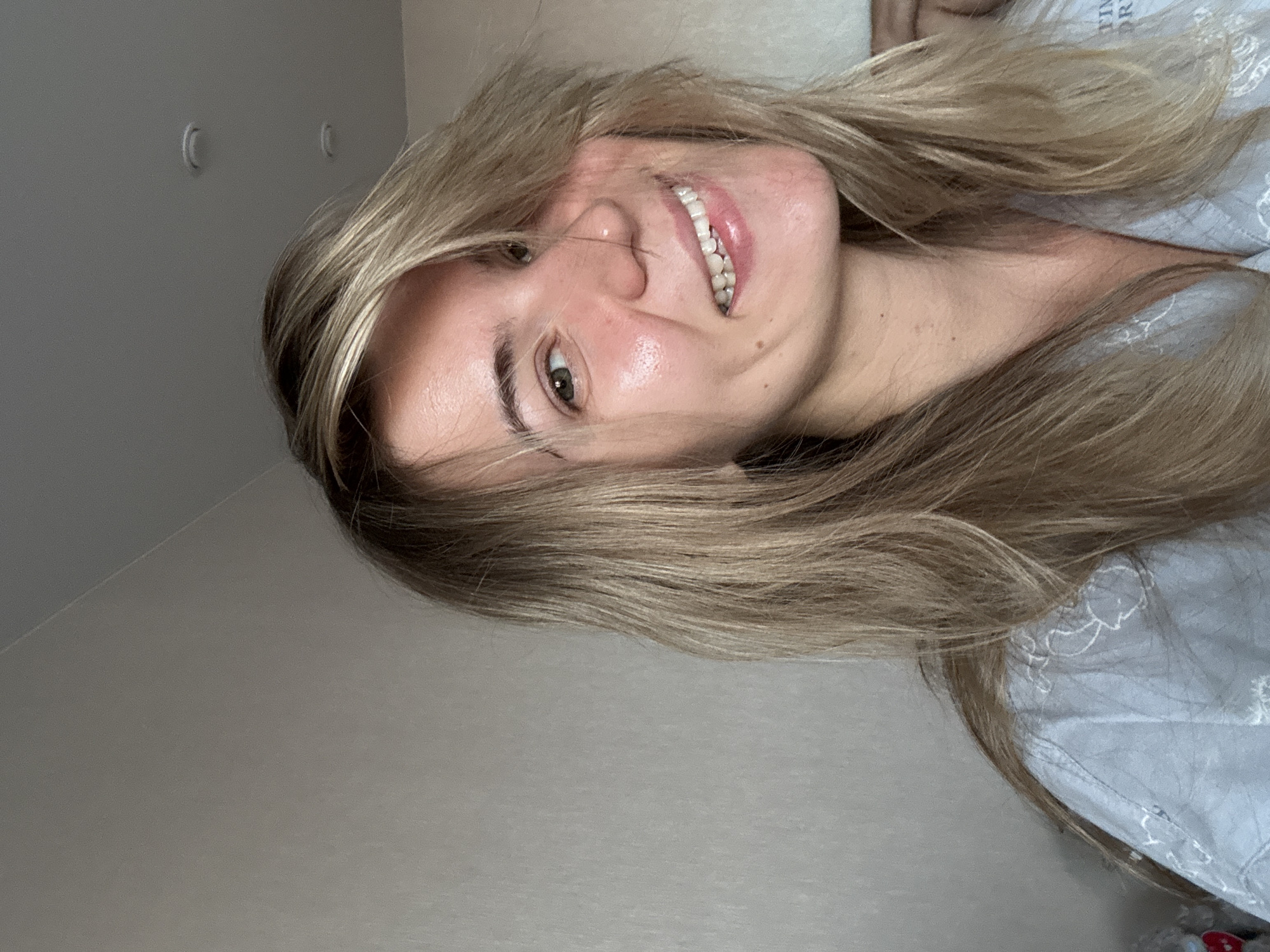 A young woman with light brown hair is smiling. She is indoors with a light gray wall in the background. The location is Unknown, Unknown.