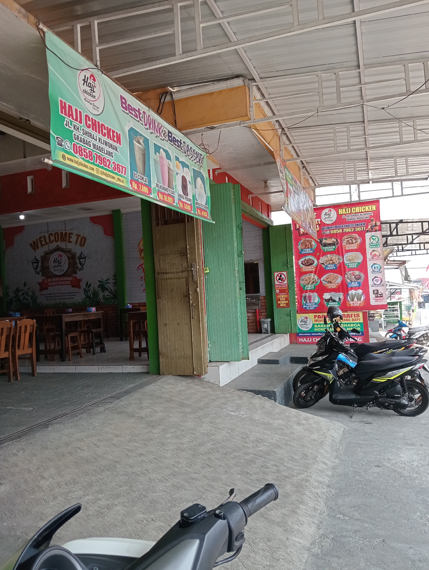 The image shows the exterior of a restaurant named "Haji Chicken" located in Grabag, Indonesia. A green banner hangs from the ceiling above the entrance, advertising "Best Drink & Best Desserts" and listing "Haji Chicken" with its address: JL. KH. SHIRAJ KLIWONAN, GRABAG MAGELANG. A phone number, 085879623677, and website, www.hajichicken.com, are also visible. Inside the restaurant, tables and chairs are set up. To the right of the entrance, a red banner displays various food items offered by Haji Chicken, including fried chicken, chicken steak, chicken burger, and French fries. Several motorcycles are parked in front of the restaurant. The overall scene depicts a commercial establishment in a public area.