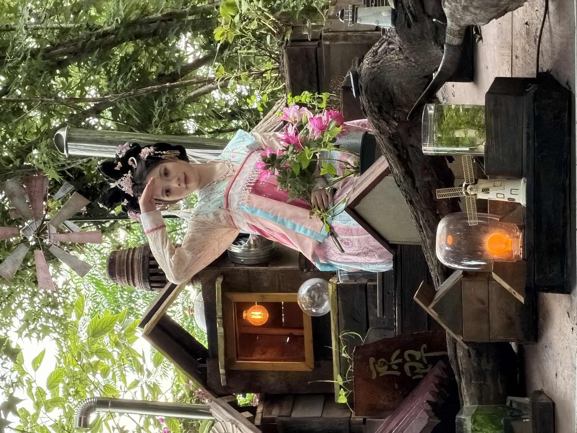 A young woman with dark hair styled in an ornate updo adorned with pink flowers and gold accessories stands in what appears to be a meticulously crafted miniature village or diorama, set against a backdrop of lush green foliage and trees, suggesting an outdoor or garden setting. She is wearing a traditional-style garment with pastel pink, blue, and white layers, embellished with delicate beadwork around the neckline. Her right hand is raised to her forehead as if shielding her eyes from the sun or in a gesture of greeting. In her left hand, she holds a bouquet of bright pink flowers.
The diorama itself is intricate, featuring small wooden structures resembling houses with pitched roofs, a prominent window with an illuminated orange Edison-style bulb, and a glass sphere on a shelf. To the left of the woman, a metal pipe extends upwards, connected to a weathered, possibly rusted, windmill structure. Further elements of the diorama include what looks like a piece of wood with large, calligraphic characters inscribed on it, various small buildings, a miniature lighthouse, and a glass container holding green aquatic plants. The overall aesthetic of the diorama appears to be a whimsical, perhaps steampunk-inspired, village. The lighting suggests it is daytime, with dappled sunlight filtering through the leaves. The Yuzhong District of China is known for its hilly terrain and unique cityscape, though the specific location within the district is not determinable from the image alone.