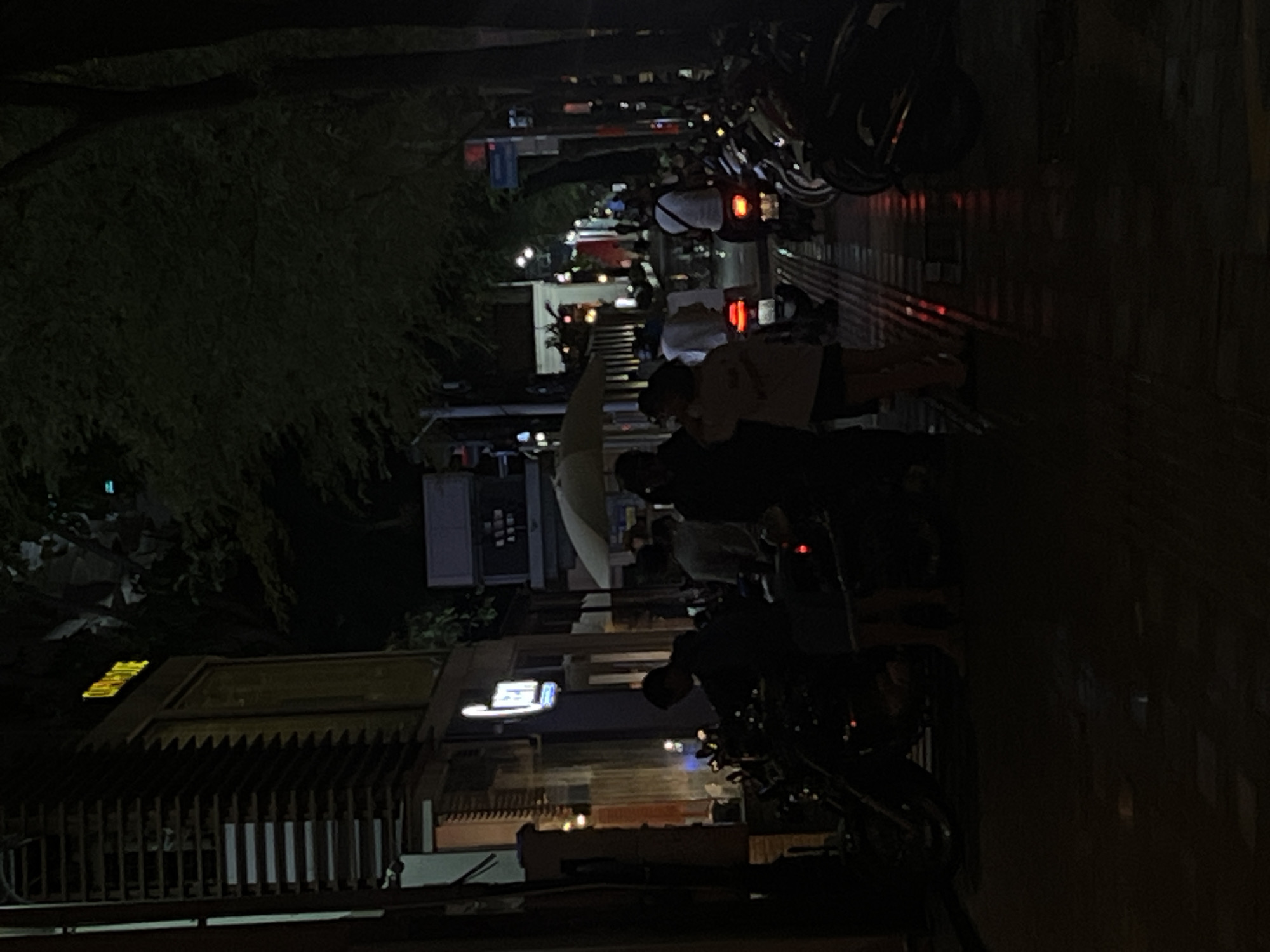 This image depicts a street scene at night in Thủ Đức, Vietnam. The foreground is dominated by parked motorcycles, with several people standing near them. A woman in a light-colored top and dark shorts is visible in the center, facing away from the camera. To her left, a man in a dark shirt is leaning over a motorcycle. Further down the street, more motorcycles are parked along the sidewalk, and several individuals are walking or standing.
The buildings lining the street are dimly lit. On the left, a building with a striped facade features illuminated signs. One sign in yellow lettering reads "CAROUSE". Another sign above a doorway is a white circular logo with blue lettering. Trees with dense foliage overhang the street, casting shadows and contributing to the dark ambiance. The street surface appears wet, reflecting the ambient light. Traffic lights and streetlights are visible in the distance, illuminating parts of the street and sidewalk. The overall scene suggests a busy urban area at night, likely a commercial or residential street with ample motorcycle parking.