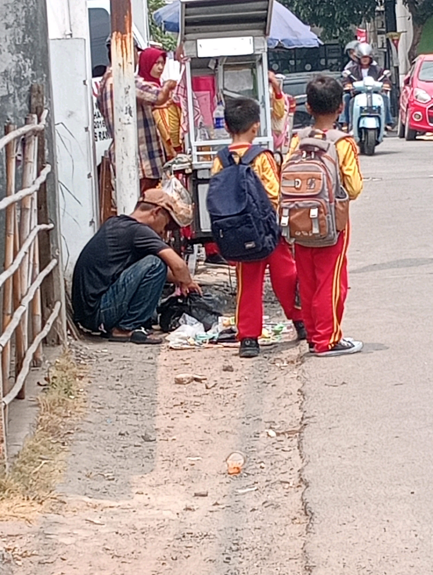 In an urban setting in Bandar Lampung, Indonesia, a man wearing a black t-shirt and blue jeans crouches on a dirt pathway next to a building. He is sorting through a pile of black garbage bags, his head bowed. To his right, two young boys, presumably students, stand facing away from the camera. They are both wearing matching red and yellow uniforms with backpacks. The boy on the left has a dark blue backpack, while the boy on the right has a brown and grey backpack. Behind the children, a light blue scooter with a rider wearing a helmet is moving on the asphalt road. To the far right of the image, the rear of a red car is visible. A vendor's cart with a white canopy is positioned between the children and the street, with a woman in a red hijab and checkered shirt visible behind it. A wooden fence runs along the left side of the pathway. The ground is a mix of dirt, gravel, and scattered debris.