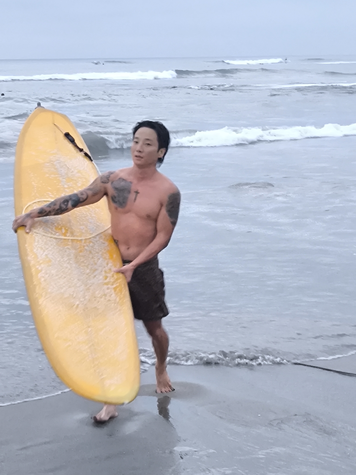 A shirtless Asian man with tattoos stands on a sandy beach, holding a yellow surfboard. He has dark hair and is wearing dark shorts. The surfboard is positioned vertically in front of him. The ocean stretches behind him with small waves breaking in the distance. The beach appears wet and grayish-brown. The scene suggests a cloudy day. The location appears to be a beach in Canggu, Indonesia.
