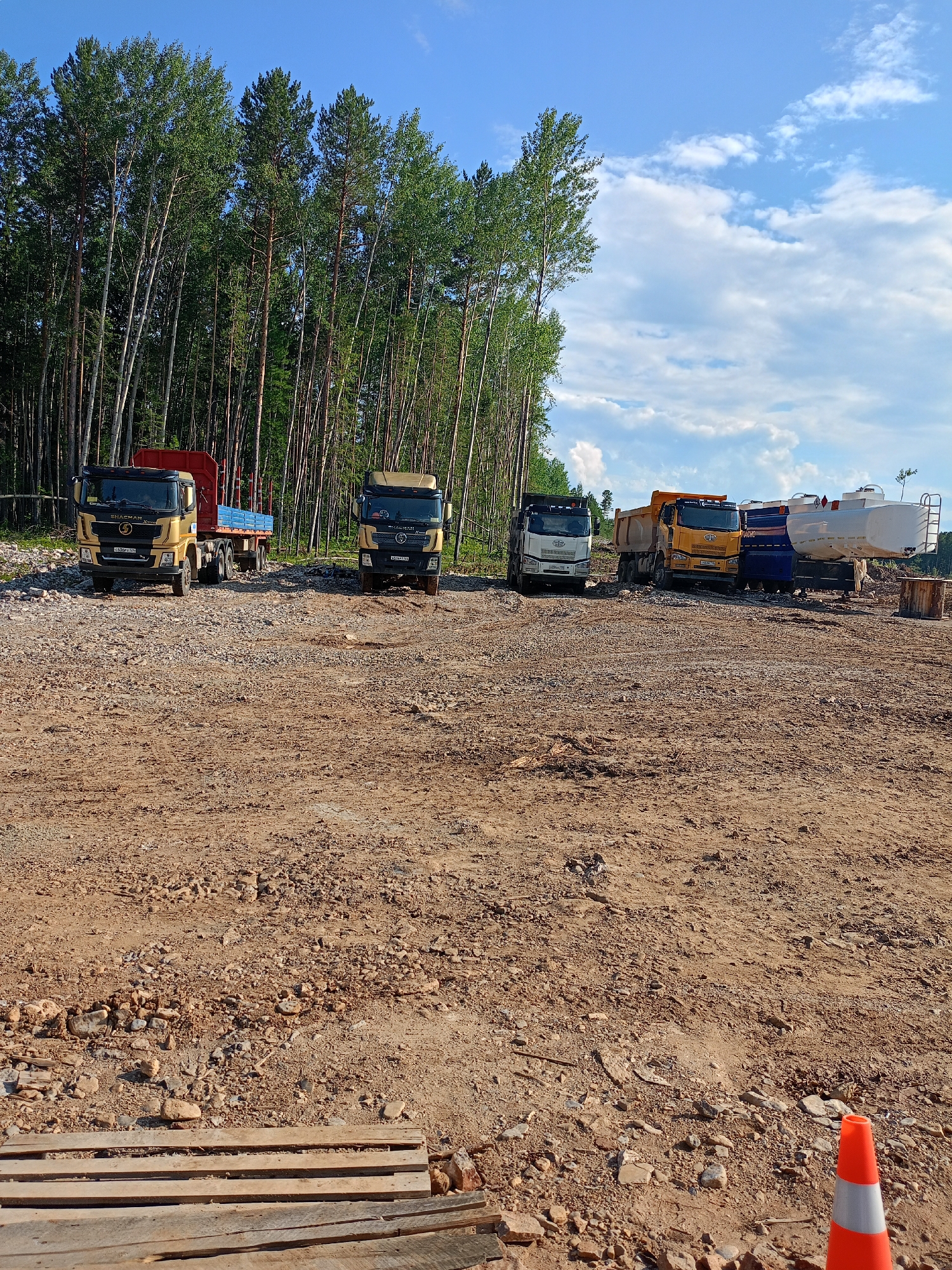 In Vitim, Russia, a construction site is depicted under a bright blue sky with scattered white clouds. The foreground is dominated by a rough, unpaved surface of dirt and gravel, with discarded wooden pallets and an orange traffic cone visible. In the mid-ground, a line of several trucks are parked. From left to right, the first truck is gold with a red trailer, followed by another gold truck. Next are two yellow dump trucks, one appearing to be hauling material, and further to the right, a white tanker truck is parked alongside a blue tanker truck. Behind the trucks, a dense line of tall, slender trees, primarily birches and pines, forms a natural backdrop. The overall scene suggests an active or recently active construction area in a forested environment.