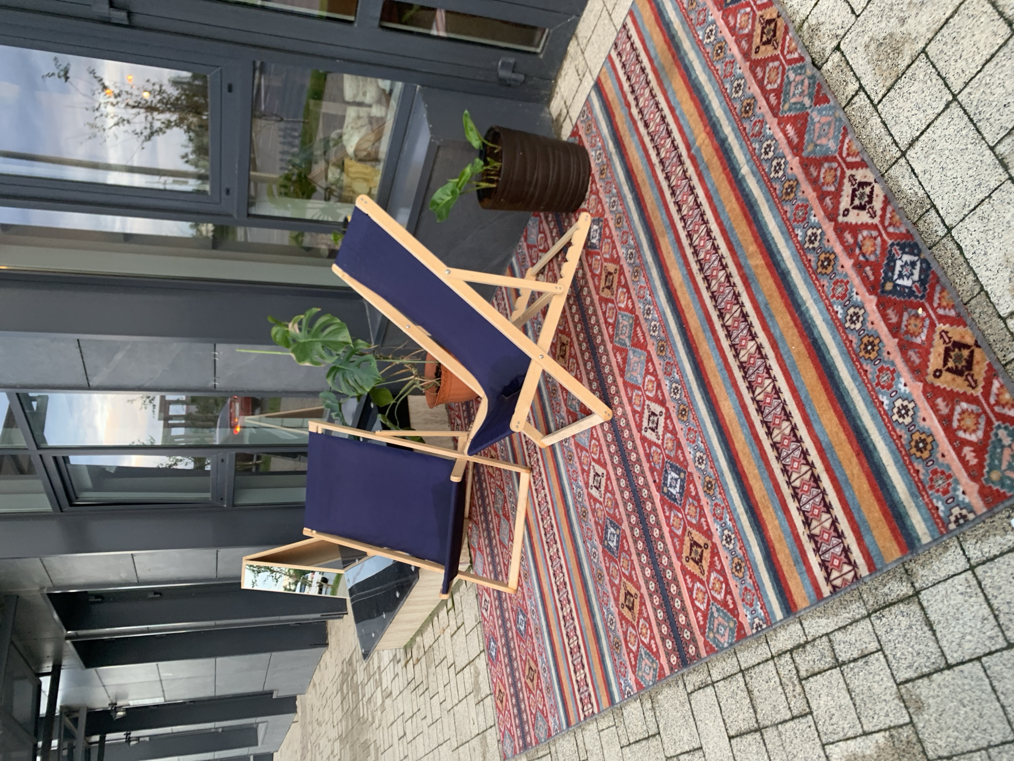 The image depicts an outdoor seating area situated on a tiled patio in front of a modern building with large glass windows. Two dark blue canvas deck chairs with wooden frames are positioned on a vibrantly patterned rug. The rug features horizontal stripes in red, orange, yellow, blue, and white, with intricate geometric motifs. A tall, slender mirror on an A-frame stand is placed to the left of the chairs, reflecting the building's exterior. In the background, potted plants, including a Monstera, add greenery to the scene. The overall impression is one of a stylish and inviting outdoor space, potentially part of a commercial establishment or a well-appointed residence. The building's architecture, with its dark grey facade and large glass panels, suggests a contemporary urban setting. The reflection in the windows indicates an overcast sky and hints at surrounding trees and structures, suggesting the location is within a city. While the specific location is stated to be in Stavropol, Russia, this cannot be definitively confirmed from the visual information alone. However, the aesthetic and architectural style are consistent with urban environments globally. There are no people visible in the image.