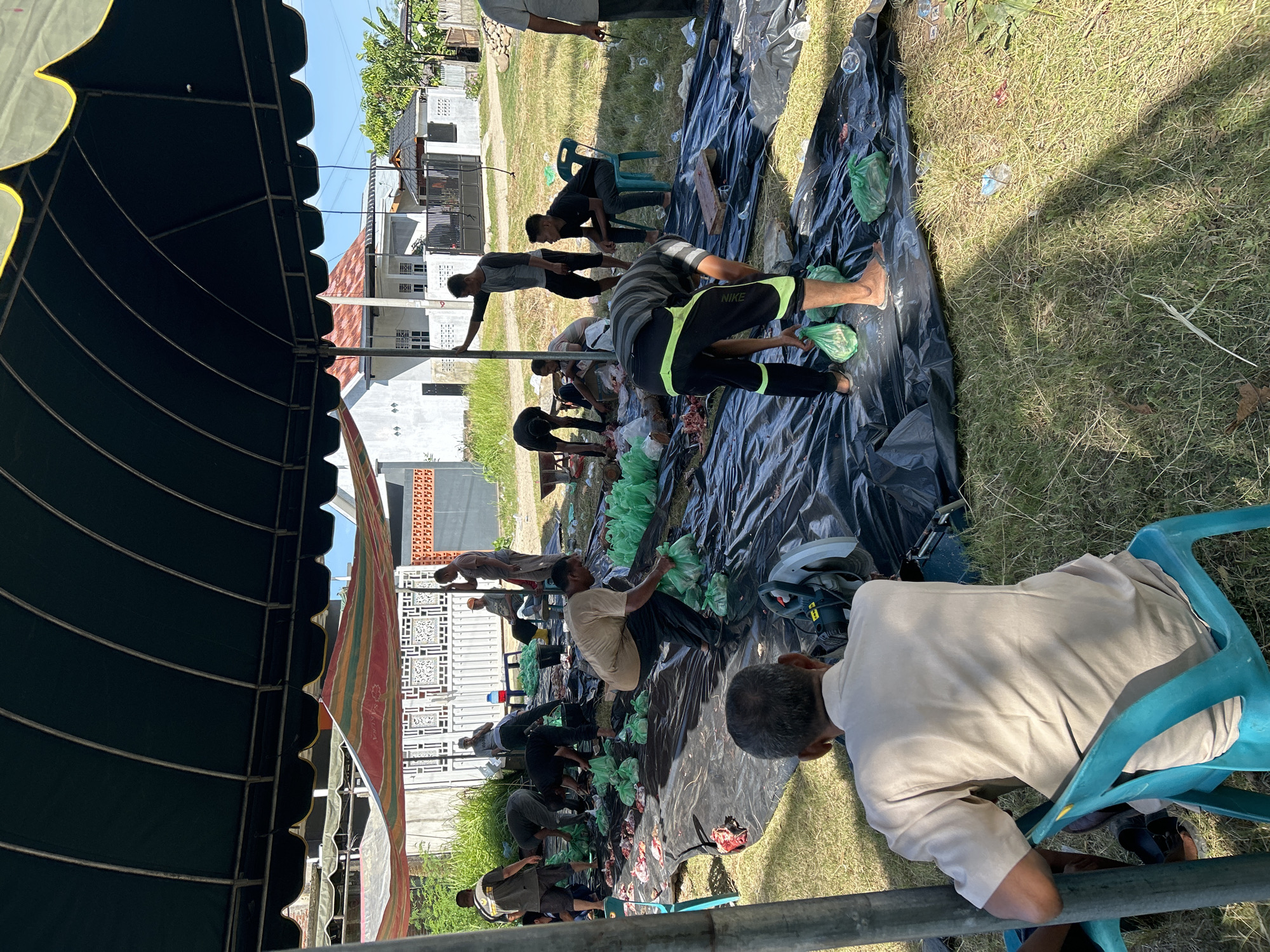 A group of people is gathered outdoors in Lhokseumawe, Indonesia, under a tent. They are seated on chairs and on the ground, and are working with what appear to be bags and possibly raw meat spread out on a large sheet of black plastic. A power saw is also visible near the plastic sheet. There are houses in the background and grass under the tent.