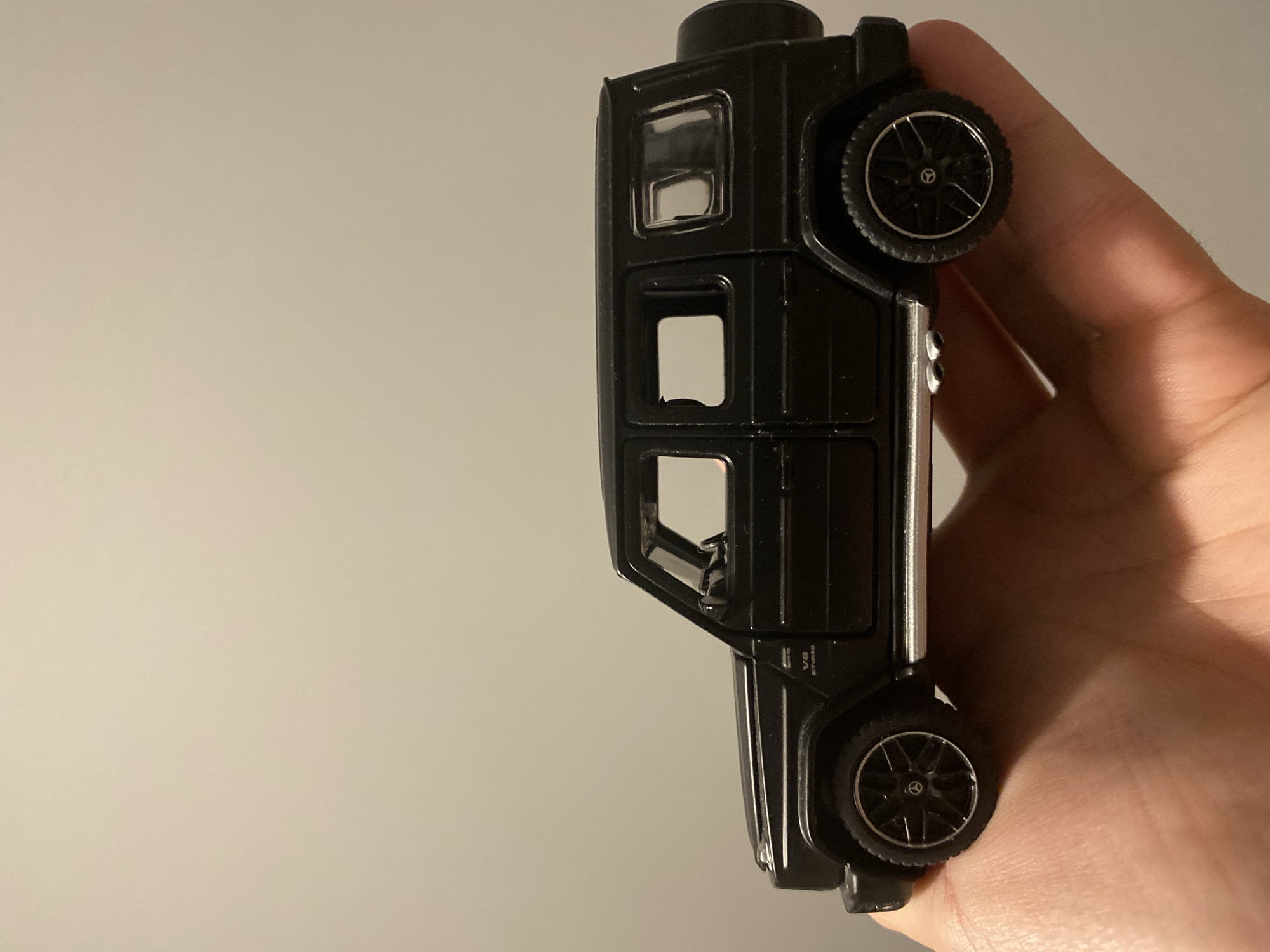 A black Mercedes-Benz G-Class toy car is held in a person's hand. The car is black with silver trim along the bottom and exhaust pipes. The wheels are black with silver accents and feature the Mercedes-Benz logo. The car has a spare tire mounted on the back. The background is a plain, light-colored wall. There is no information in the image to suggest a location in Łódź, Poland.