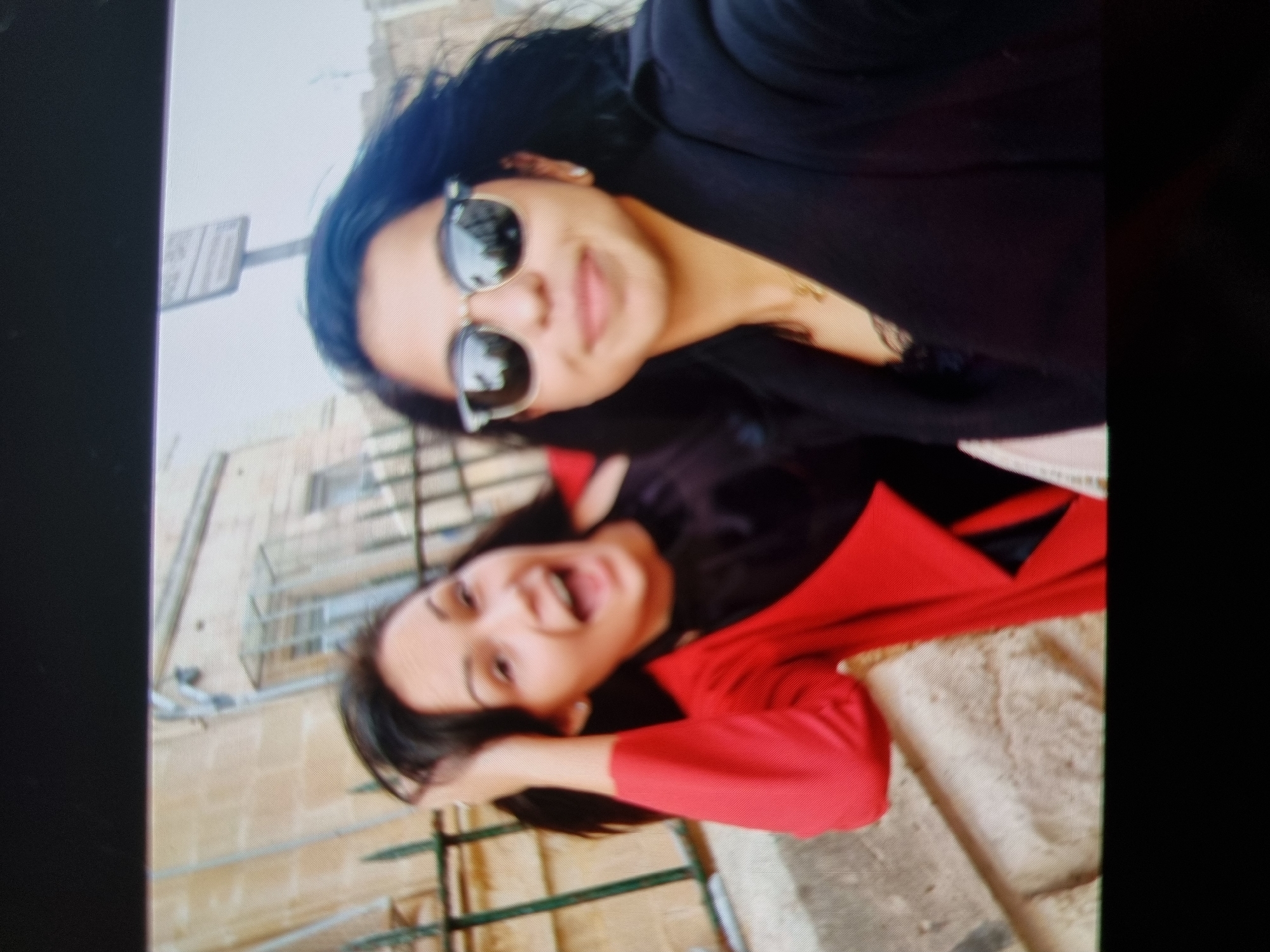 Two women are posing for a photo outdoors. The woman at the top is wearing sunglasses and a black blazer. The woman at the bottom is wearing a red shirt and smiling. They are standing next to a wrought iron fence with a stone wall in the background. Based on the architectural style and materials, this could be in the Old Town area of Plovdiv, Bulgaria. A sign is visible on the left side, though the text is unreadable. The ground appears to be paved.