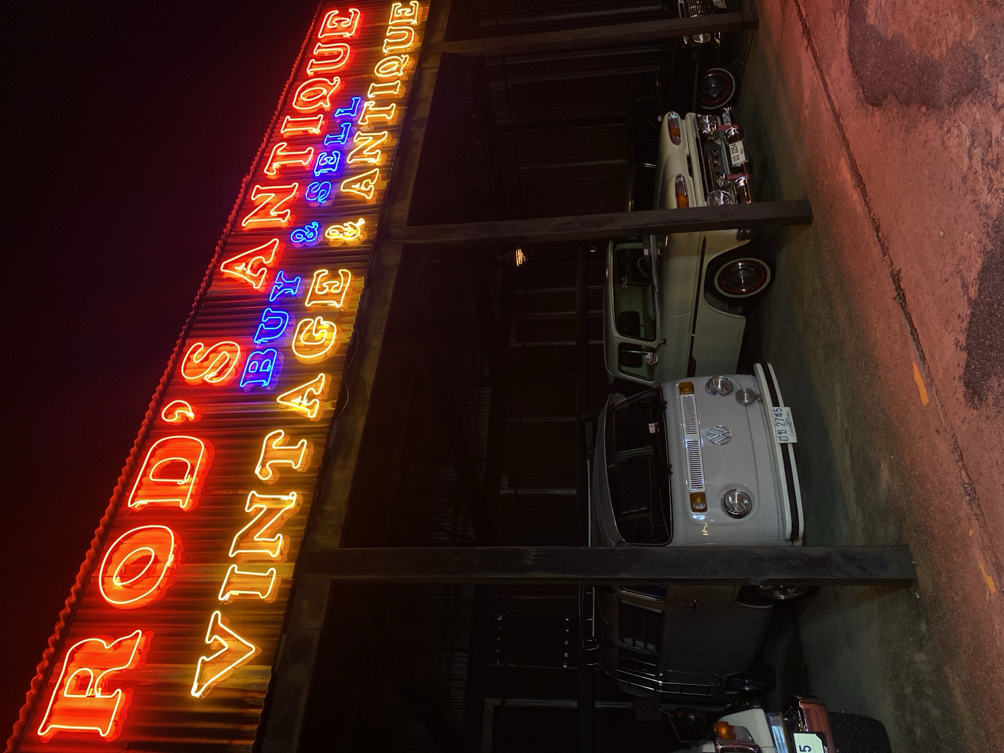 The image depicts "Rod's Vintage Antique" car dealership, presumably in Bangkok, Thailand. A large neon sign with the name of the business is visible, in red, yellow, and blue. Below the sign are several classic cars parked under a dark shelter, including a white Volkswagen van. The license plate "ฮข 2745" is visible on the van, indicating Thailand. Another license plate, "บบ 2254" is also visible on a lighter-colored vehicle. The ground appears to be concrete with yellow markings. The image is dark, suggesting it was taken at night.