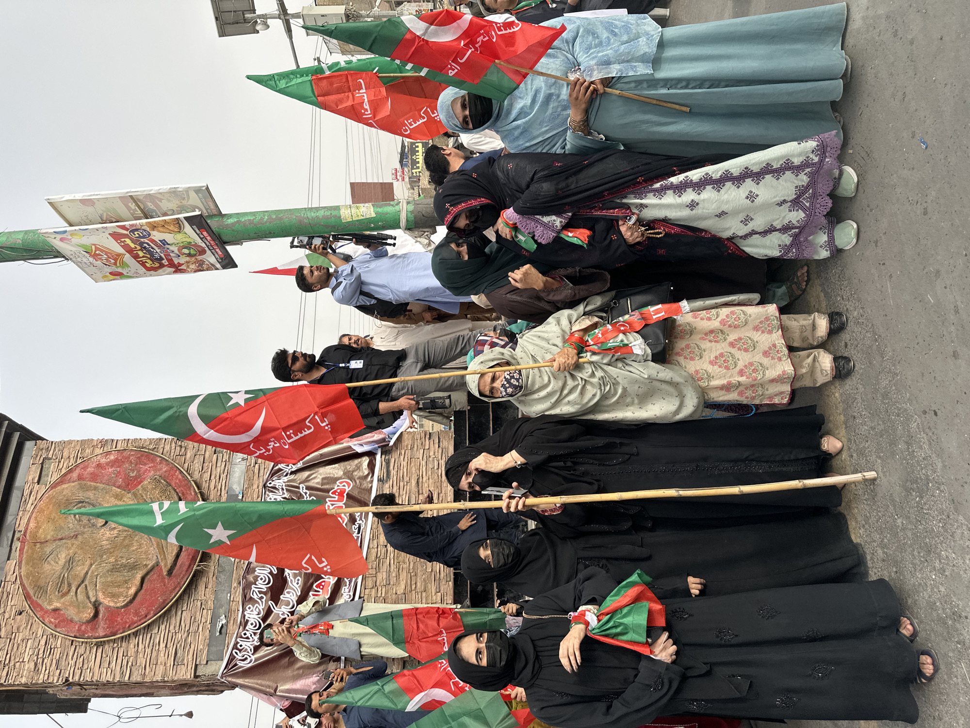 In Quetta City Tehsil, Pakistan, a group of individuals, predominantly women, are gathered, holding political flags. The flags are red and green, featuring a crescent moon and star, consistent with the flag of Pakistan, and also other flags with red and green sections and a white star, possibly representing a political party. The people are dressed in traditional attire, with many women wearing hijabs and abayas. One woman in the foreground is holding a small red and green flag. Behind them, a large circular relief of a man's face is visible on a textured stone structure. Several other individuals, including men, are visible in the background, some holding cameras and phones. A green lamppost with signage is also present. The setting appears to be an outdoor public space, with a paved ground surface. The overall atmosphere suggests a political gathering or rally.