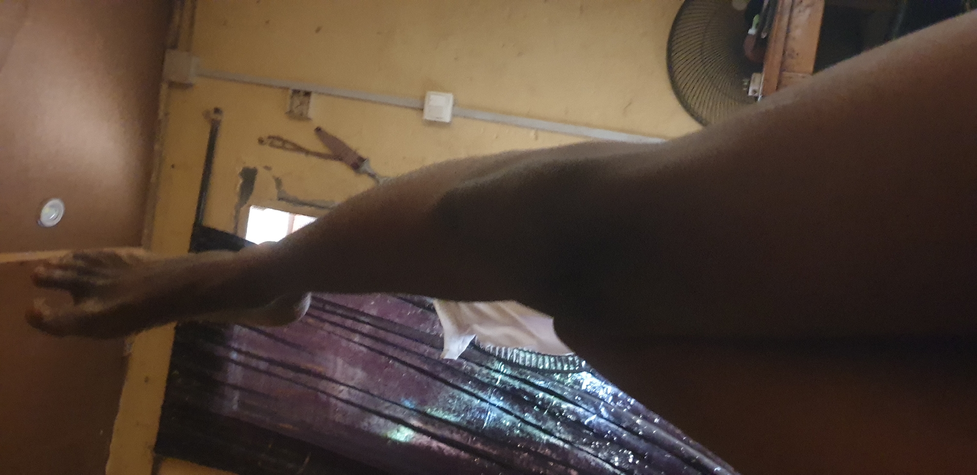 In Yelwa, Nigeria, a low-angle shot captures a person's legs and feet against a backdrop of an indoor environment. The dominant colors are warm yellows and browns from the walls and ceiling. To the left, dark, sheer curtains with a shimmering effect hang over a window, allowing some ambient light to filter through. A white sheet or piece of fabric is visible beneath the curtains.
The ceiling features a circular light fixture. A white electrical conduit runs vertically along the wall, terminating in a white junction box. To the right, part of a wooden table is visible, on which a black oscillating fan is positioned. The overall impression is of a room with modest furnishings.