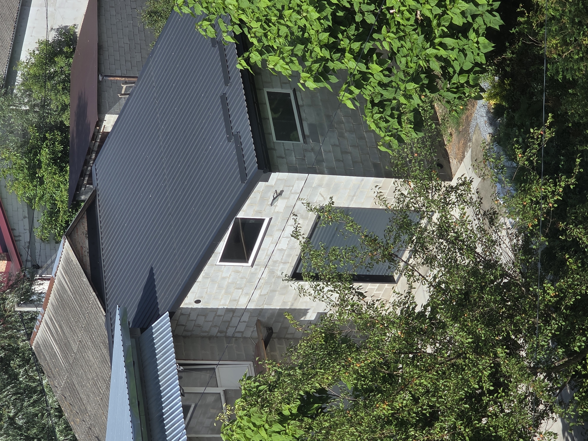 This is a drone shot, taken from a high angle, of several residential buildings and trees in the city of Dubno, Ukraine. The buildings are constructed from concrete blocks and corrugated metal, with some roofs made of slate. There are various trees with green foliage and some with fruit visible. The overall setting appears to be a residential area with a mix of older and newer construction.