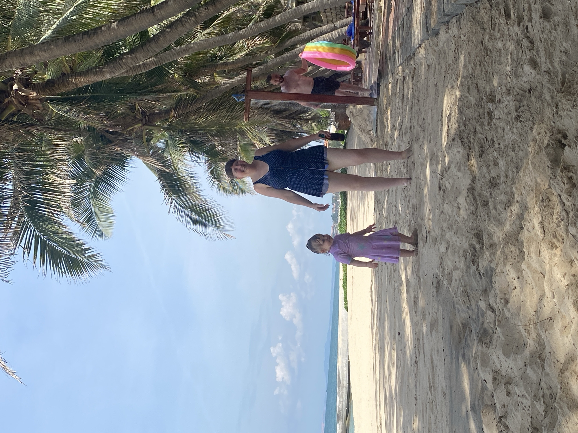 The image shows a beach scene in Phu Thuy Ward, Vietnam, on a clear, sunny day. The foreground is dominated by palm trees with lush green fronds casting shadows on the sandy beach. In the middle ground, a woman in a dark blue patterned swimsuit stands on the sand, holding a phone in her right hand. To her right and slightly further back, a young girl in a purple dress stands facing the sea. In the background, a man is visible holding a colorful inflatable toy. The ocean stretches out to the horizon, with a thin line of clouds in the distant sky. The overall atmosphere is relaxed and summery, suggesting a family outing at the beach.