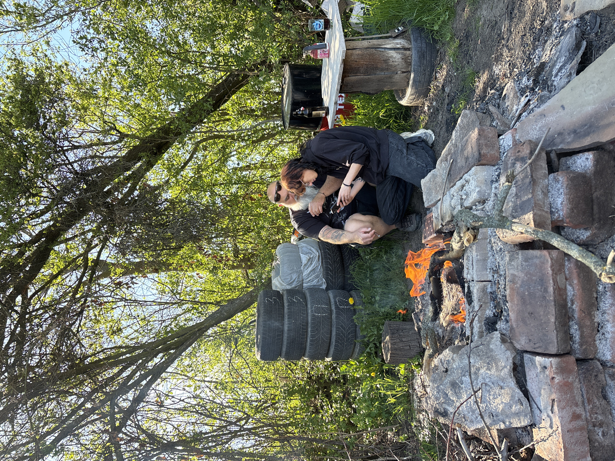 This outdoor image, captured in Verinsko, Bulgaria, depicts a casual gathering on a bright, sunny day. The scene features two individuals, a man and a woman, in close proximity next to a lit campfire. The man, with a long white beard and sunglasses, is wearing a dark t-shirt, revealing tattoos on his arm. The woman, with dark hair, is dressed in a dark hoodie and jeans, leaning into the man in what appears to be an affectionate or intimate embrace.
They are positioned on a dirt and grassy patch of ground, near a makeshift fire pit constructed from bricks and stones, where a fire is actively burning with bright orange flames. To their left, a stack of used car tires is visible, some neatly piled, with one tire featuring a small yellow dandelion flower. Behind them, a rustic, makeshift table or workbench, fashioned from wood and what appears to be a black barrel, holds several bottles, including what look like alcoholic beverages. Partially obscured red text, possibly "KEL," can be seen on the side of this table structure.
The setting is an open-air environment, characterized by abundant green foliage and dense tree canopies overhead, suggesting a rural or semi-rural location. Bright sunlight filters through the leaves, indicating a late afternoon or early evening time. The overall impression is one of relaxed companionship in a natural, informal setting.