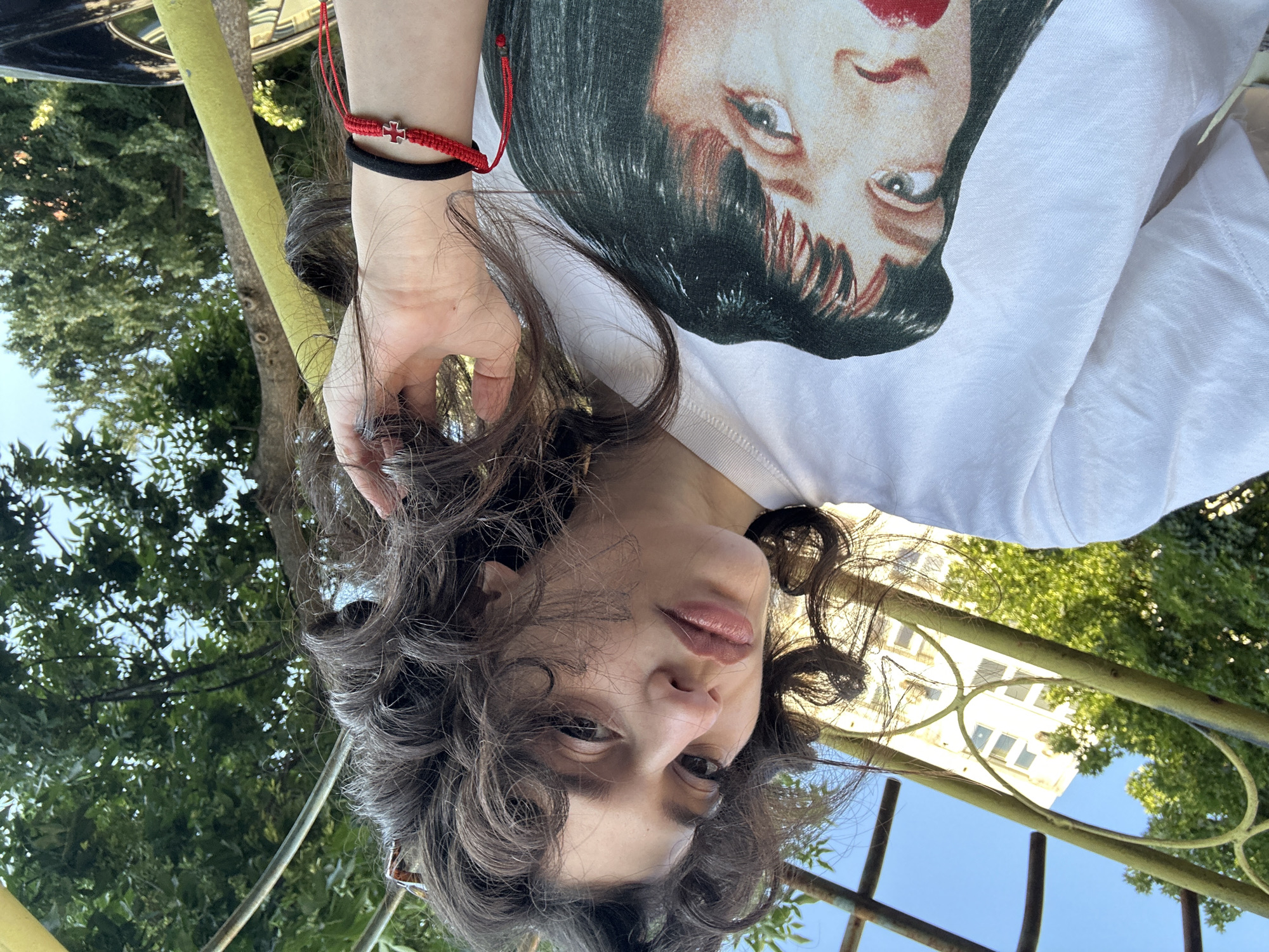 A young woman with curly dark hair is taking a selfie outdoors. She is wearing a white t-shirt with a graphic print of a woman's face on it. She has a red and black bracelet on her right wrist. In the background, there is a building with multiple windows and trees. The setting appears to be a park or courtyard, possibly in Sofia, Bulgaria, given the architectural style of the building and the surrounding foliage. The woman is holding her hair back with her hand. The lighting suggests it is daytime.