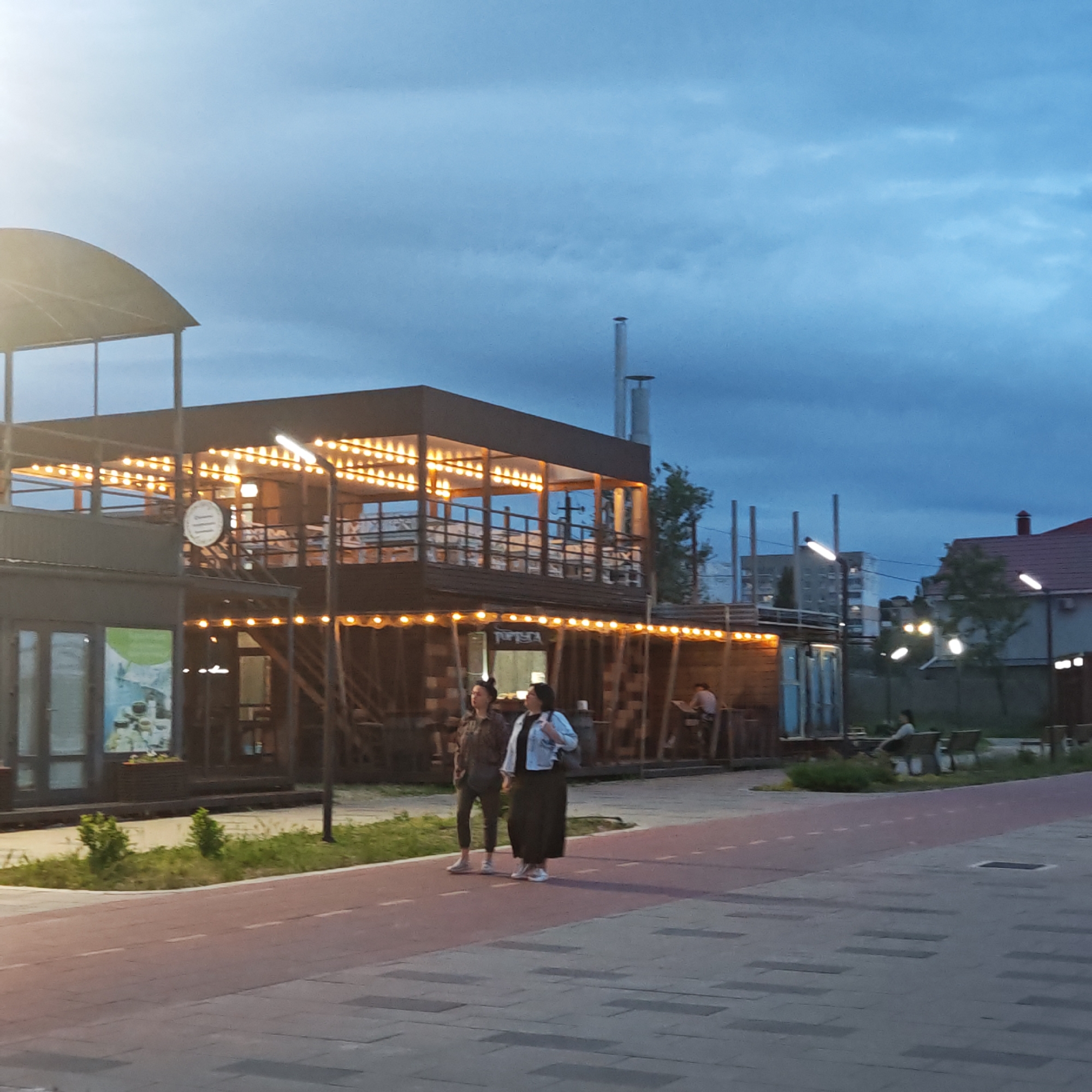 The image captures an evening scene in городское поселение Щелкино, Ukraine, characterized by the warm glow of artificial lights against a twilight sky. The sky transitions from a lighter, slightly hazy blue on the left, possibly indicating a setting sun or strong artificial light source just out of frame, to a deeper, cloud-streaked blue towards the right.
In the foreground, two women walk on a paved area, which features a distinct reddish-brown path with dashed white lines, likely a designated pedestrian or cycling lane, adjacent to a grey sidewalk. The woman on the left wears dark clothing, while the woman on the right wears a light-colored jacket over a dark skirt or dress and carries a bag.
The central focus is a two-story building, possibly a cafe or restaurant, adorned with numerous string lights that cast a warm, inviting glow. The upper level features an open balcony area. A circular sign hanging from this building is partially visible, bearing the text "Сладкие Воспоминания" (Sweet Memories). To the left of this main building, there's another structure with large windows, one displaying a poster with an astronaut image and some products. Small bushes and grass line the edge of the paved area.
Further in the background, to the right, another person is seen sitting at an outdoor table. Modern streetlights with elongated fixtures are illuminated, contributing to the overall evening ambiance. Distant buildings, including a house with a red roof and some apartment blocks, are visible against the darkening sky, indicating an urban or semi-urban setting. The overall mood is calm and relaxed, suggesting leisure activity during the evening hours.