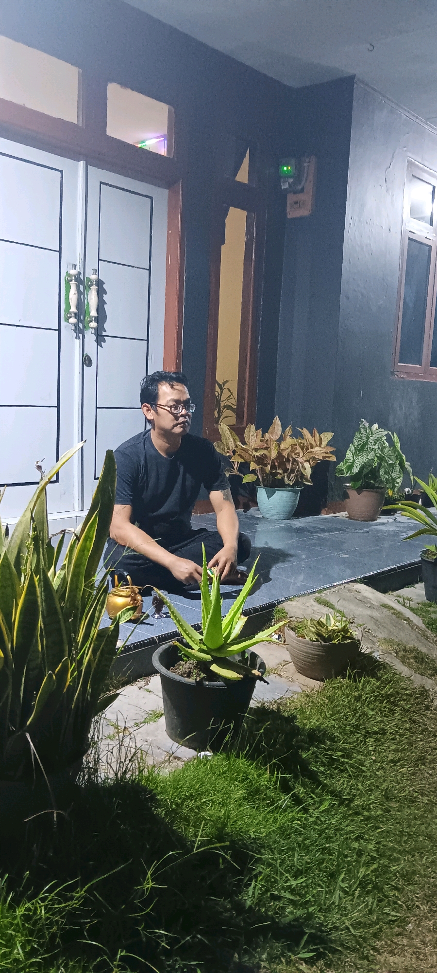 A man wearing a black t-shirt and black pants sits cross-legged on a tiled porch in front of a house at night. He wears glasses and has short, dark hair. To his left is a golden teapot and a small glass. In front of him is a potted aloe vera plant and to his left, a potted snake plant. Behind the man are several other potted plants. The porch is dark, with light coming from the house and a security camera visible on the wall above a door. The background shows a dark, grassy area.