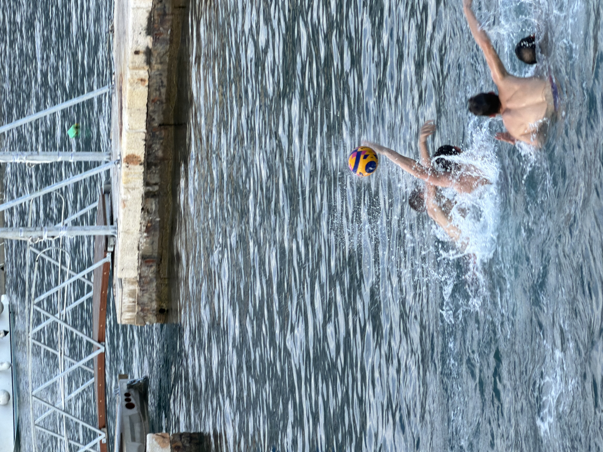 The image depicts a water polo game taking place in the Adriatic Sea, near the marina in Budva, Montenegro. Several individuals are in the water, actively engaged in the sport. One person is in the process of throwing a water polo ball, which is visible in mid-air with its distinct yellow, blue, and orange panels. Splashes of water indicate the dynamic nature of the game. In the background, parts of the marina infrastructure are visible, including a pier with concrete blocks and a metal ramp or walkway. The water is a deep blue, reflecting the sky.
