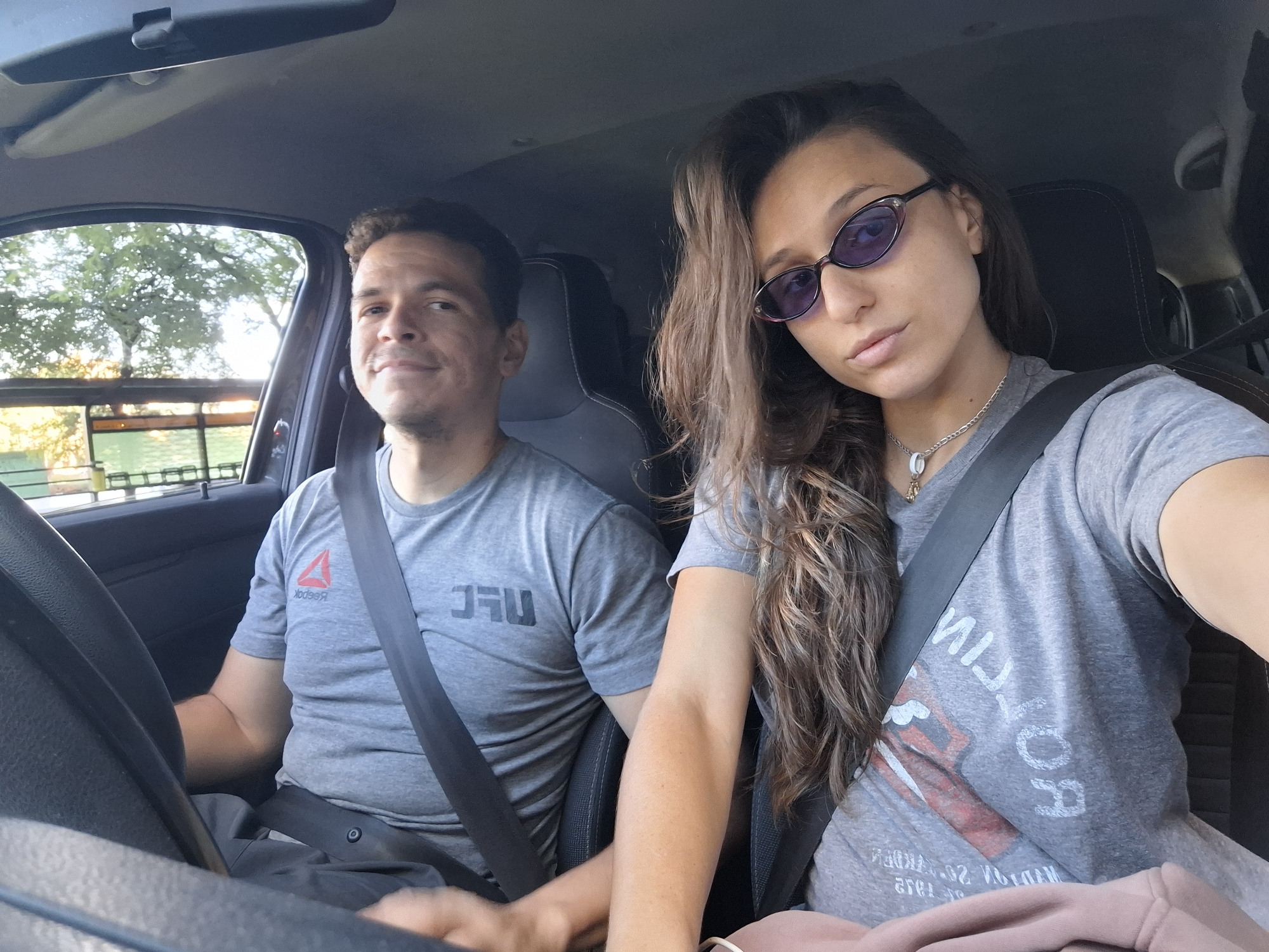 Inside a car, a man and a woman are posing for a selfie. The man, in the driver's seat, wears a grey t-shirt with a UFC logo and a seatbelt. The woman, sitting next to him, is also wearing a seatbelt and a grey t-shirt, along with purple sunglasses. She has long brown hair. The location appears to be in the city of Buenos Aires, Argentina, as suggested by the appearance of the street and possibly trees visible through the car window.
