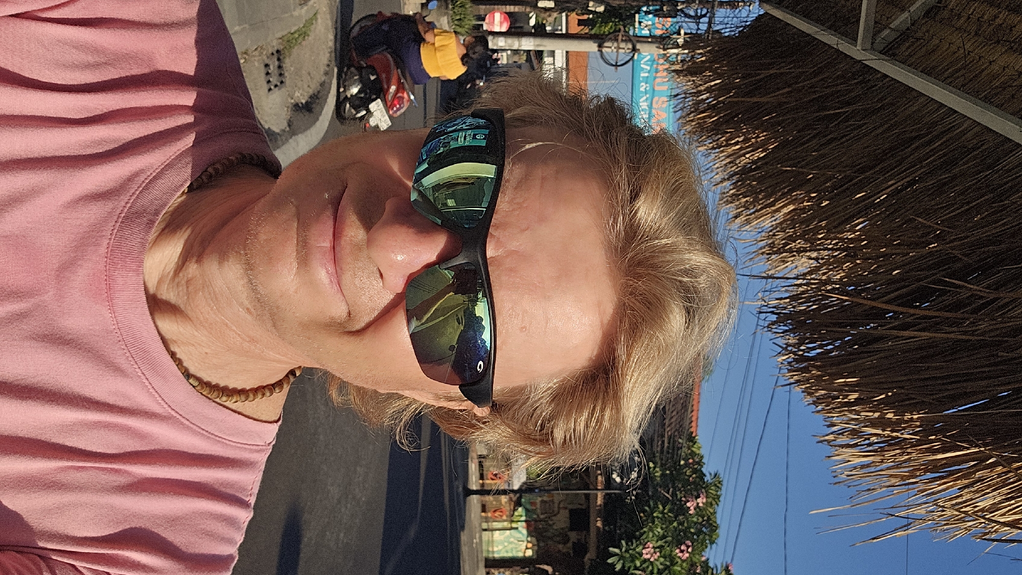 The image is a selfie taken outdoors, likely in Denpasar, Indonesia. The subject is a man with blond hair, wearing a pink t-shirt and sunglasses. The sunglasses reflect a street scene, including buildings, trees, and a motorbike with a rider. The background shows a street with buildings, trees, and power lines. A thatched roof structure is visible overhead.