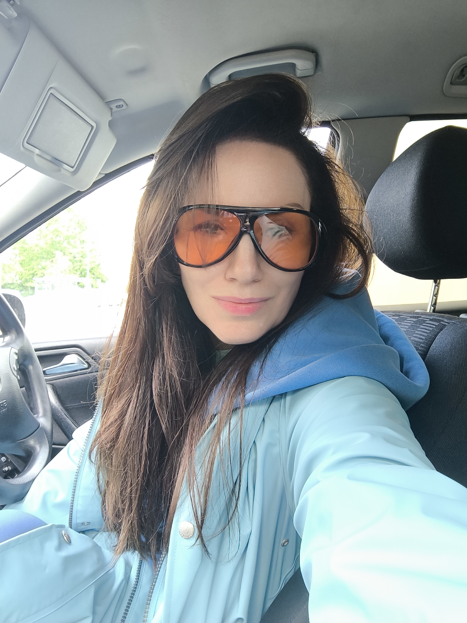 A medium shot captures a woman inside a vehicle. She has long, dark brown hair, with some strands styled in a partial bun on top of her head. She is wearing large, black-framed sunglasses with orange lenses and a light blue hooded jacket over a blue hoodie.
The interior of the car is visible, featuring a black steering wheel, a partially open sun visor, and a black headrest. The presence of a steering wheel and headrest suggests the woman is in the driver's seat.
While the car's interior details are evident, the exterior is less discernible, but judging by car's registration plate reflections on the sunglasses, the vehicle is in Latvia. The green foliage visible through the windshield implies the presence of trees, typical of Riga's landscape. The blurred objects visible in the reflection of the sunglasses are difficult to make out, there is a possibility that the car is located near a public service, or office building.