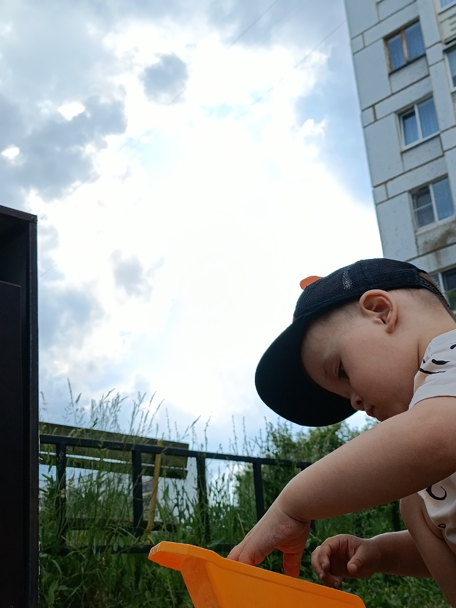 The image shows a close-up view of a young boy wearing a black baseball cap and a white t-shirt, looking down and playing with an orange object, possibly a toy. He is in an outdoor environment with lush green grass and some foliage in the foreground, suggesting a park or backyard setting. In the background, a multi-story building with windows is visible, and the sky is filled with clouds. The overall impression is that of a candid moment of a child engaged in play. The location is identified as Chaikovsky, Russia, implying this scene is taking place within the city's urban landscape.