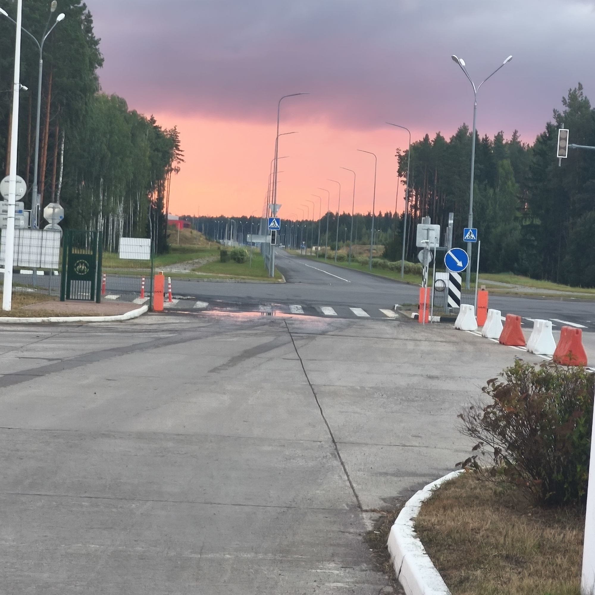 The image depicts a road at dusk in the vicinity of Драчкаўскі сельскі Савет, Belarus. The sky is a vibrant gradient of pink and purple, indicating sunset. The road is paved and appears to be a multi-lane highway or a large access road, lined with numerous tall streetlights on both sides. In the foreground, there is a crosswalk, and a few traffic cones and temporary barriers are visible, suggesting some form of road work or traffic control. To the left, a green metal gate with a crest is present, potentially marking an entrance to a facility. A line of trees forms a dense forest on the sides of the road. The overall scene conveys a sense of transition, possibly a border crossing or an entrance to a restricted area, given the presence of the gate and barriers, under a dramatic twilight sky. There are no people visible in the image.