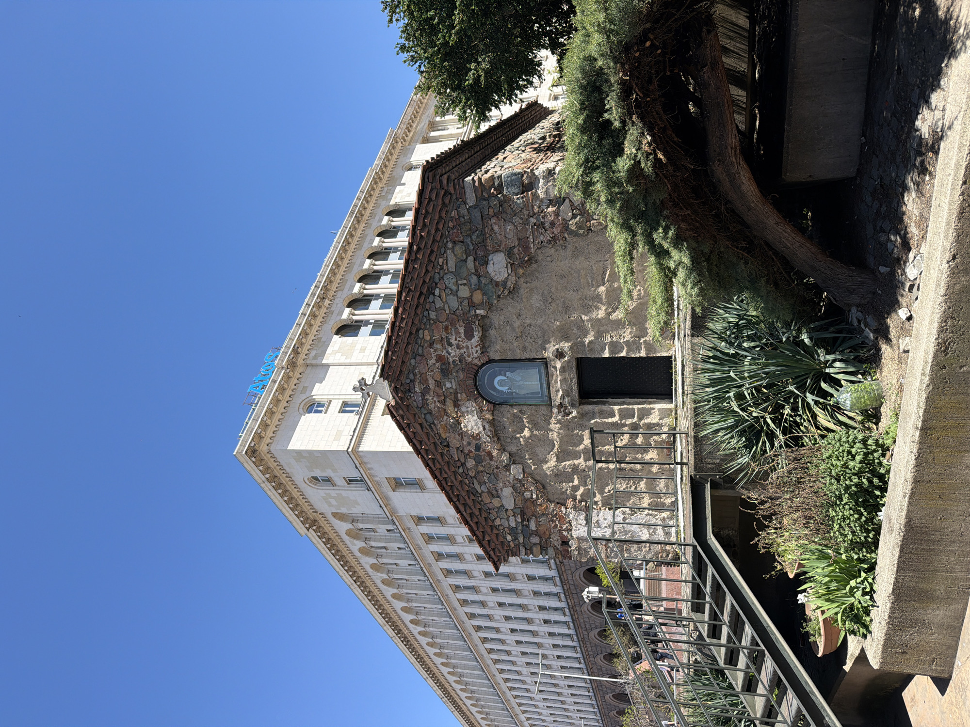 The image shows the Church of St. George Rotunda in Sofia, Bulgaria, positioned next to a larger, modern building. The rotunda exhibits a stone and brick exterior, accessible by a metal staircase. There is greenery near the base of the rotunda. The modern building is white with many windows. The sky is clear and blue.