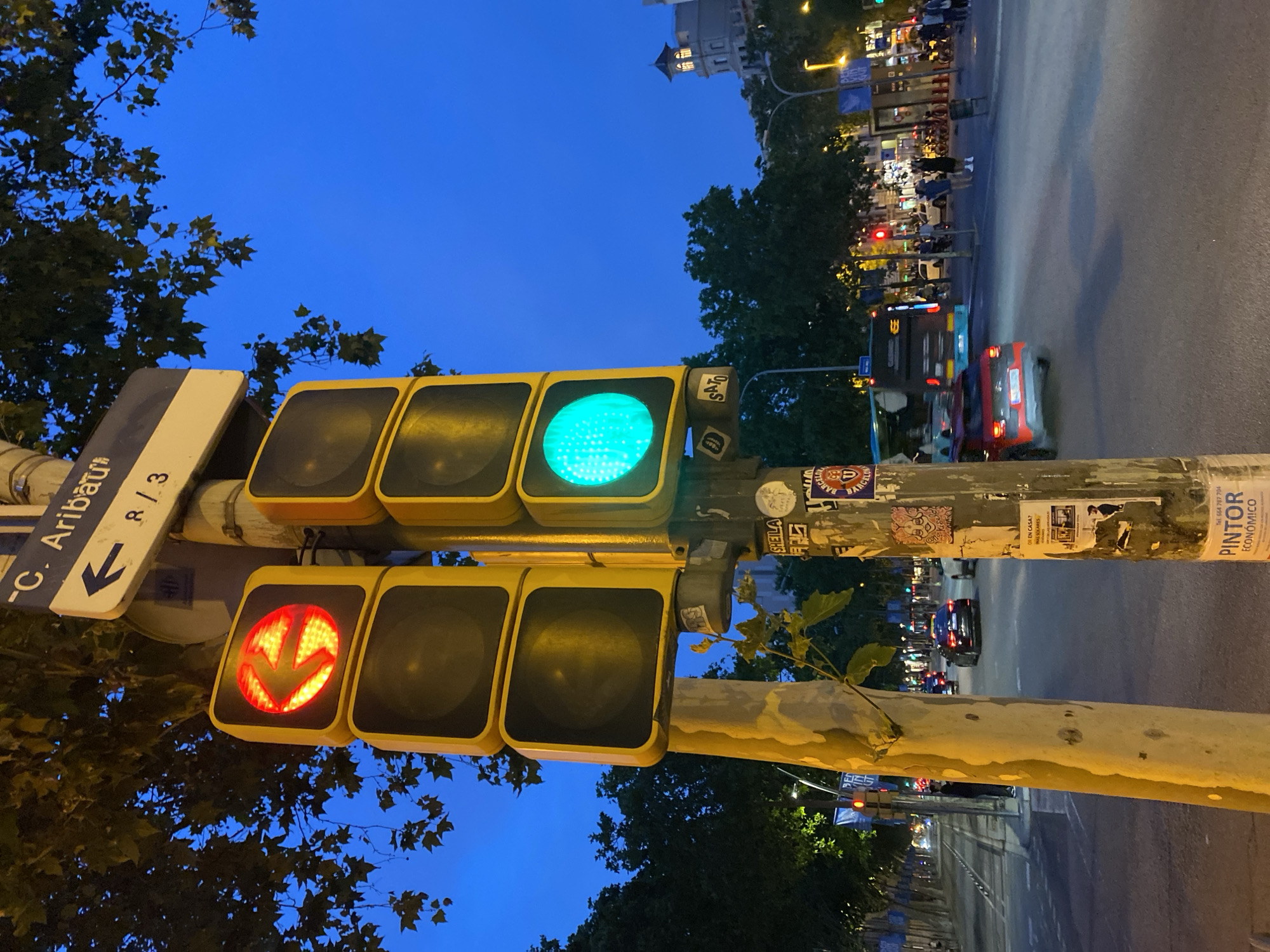 This image captures an urban street scene at twilight or night on C. Aribau in Barcelona, Spain, as indicated by the "C. Aribau" street sign prominently displayed on a pole, with an arrow pointing left and "R / 3" below it.
In the foreground, two vertical sets of traffic lights are mounted on separate poles. The left pole displays a red light with a left-pointing arrow illuminated, indicating a restriction or stop for left-turning traffic. The other two lights on this signal are dark. The right pole displays a green circular light illuminated, signifying "go" for through traffic; its other two lights are dark. The pole on the left is a lighter, rough-textured post, possibly concrete, with a small plant branch growing from its side. The pole on the right is darker, metallic, and heavily adorned with numerous stickers and posters, including one with "Barcelona" text and crest, and another for a "PINTOR ECONÓMICO."
Above the traffic lights, dark green tree foliage is visible against a deep blue twilight sky. The street extends into the background, illuminated by streetlights. Vehicles are present on the road, with a dark sedan behind the left pole and a red SUV-like vehicle in the mid-ground, both moving away from the viewer. Further down on the right, a dark-colored bus with "Galapago" text is also moving away. Distant car headlights and taillights indicate further traffic. Several pedestrians are visible on the sidewalk further down the street to the right, some standing, some walking. On the far right background, a distinctive building with a turret-like roof and illuminated windows stands out against the sky. The overall scene is one of an active city street during the evening hours.