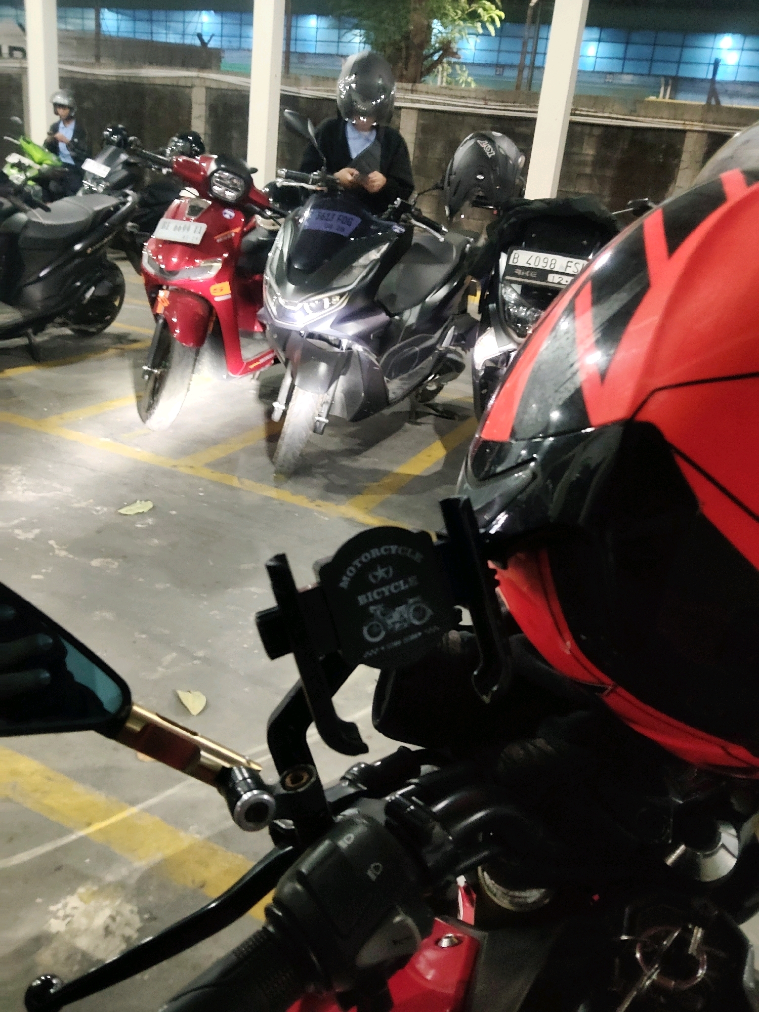The image depicts a parking lot scene with several motorcycles. In the foreground, a red and black motorcycle is partially visible, with a red helmet positioned on top. A black motorcycle with a license plate reading "B 4098 FS" is parked behind it. To the left, a red motorcycle is also present. In the background, a person wearing a helmet and a blue shirt is seated on a motorcycle. The environment appears to be an outdoor parking area, possibly within Mekarmukti, Indonesia, indicated by the type of motorcycles and license plate format common in Indonesia. The ground is paved with yellow lines marking parking spaces.