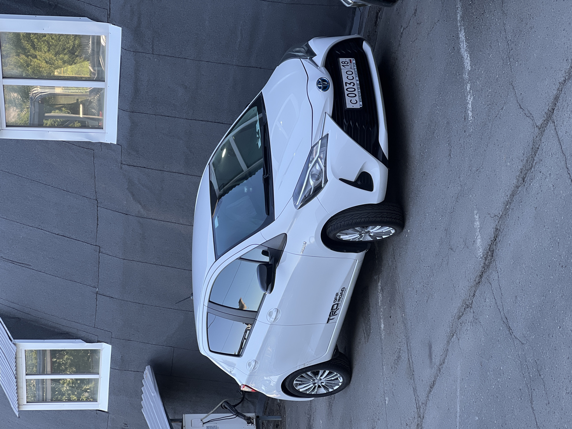 A white, third-generation Toyota Yaris (XP130 facelift) hybrid vehicle is parked on an asphalt surface with visible cracks and faded white line markings. The car faces slightly to the right of the frame. It features a Russian license plate, "C 003 CO 18 RUS," affixed to the front bumper, indicating registration in the Udmurt Republic, where Izhevsk is the capital. The license plate frame includes the text "WHENEVER YOU GO ✨ HARAT'S PUB." A "HYBRID" badge is visible on the front quarter panel, and a "TRD OFF ROAD" decal, accompanied by "TOYOTA RACING DEVELOPMENT," is applied to the lower section of the driver's side door. The car is equipped with black side mirrors and multi-spoke chrome alloy wheels.
Behind the vehicle stands a dark gray building with a distinctively textured, possibly panelled or shingled, facade that angles inward towards the top. Two white-framed dormer-style windows, each topped with a small corrugated metal awning, are set into the upper part of the building's wall, reflecting trees and the sky. To the left of the car, an external utility unit, likely an air conditioner or part of a ventilation system, is mounted on the building wall, also sheltered by a small corrugated metal cover. The scene is illuminated by natural daylight. No individuals are present in the image.