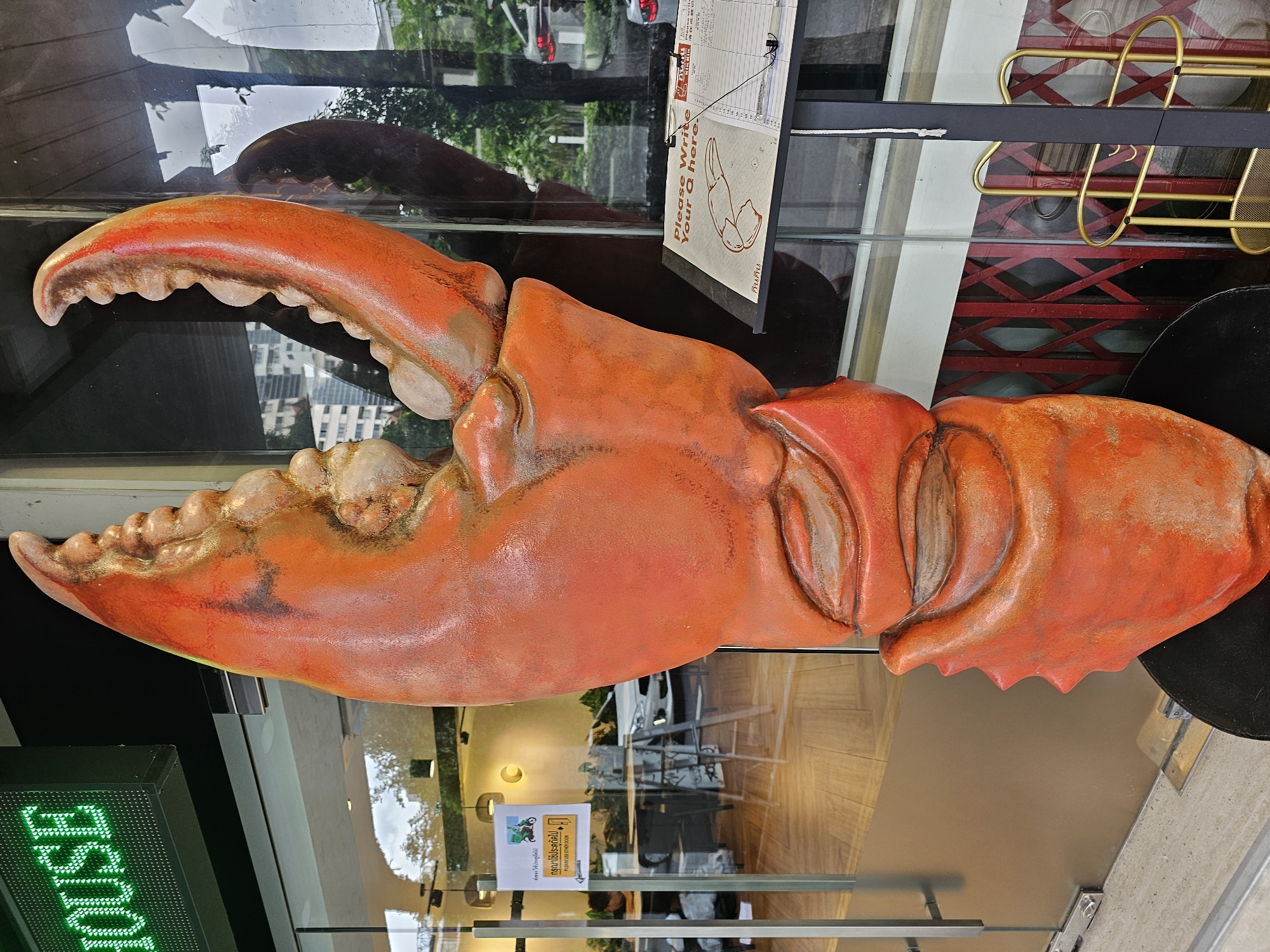 A large, orange, artificial crab claw sculpture stands prominently in front of a building entrance. The sculpture is detailed, with a textured surface resembling a real crab claw. To the left of the sculpture, a green LED sign displays the word "HOUSE" in block letters. A glass door is visible behind the sculpture, reflecting the surroundings. A yellow sign with Thai text and an illustration of a motorcycle delivery driver is affixed to the glass door. Inside the building, tables and chairs can be seen. To the right of the sculpture, a black stand holds a clipboard with a sign that reads "Please Write Your Q here." and a pad of paper with a list. The background shows a street scene with trees, buildings, and a red, accordion-style gate. The scene appears to be outdoors in a city environment.