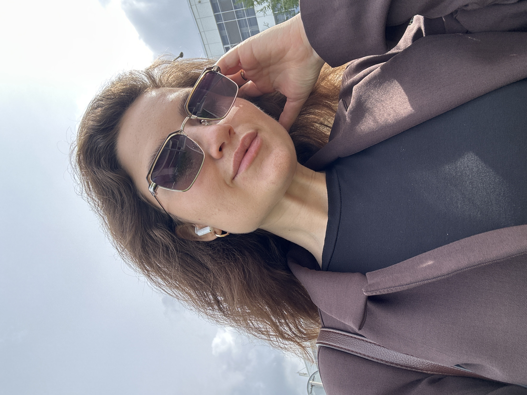 A young woman is taking a selfie outdoors, under a partly cloudy sky. She is wearing a brown blazer over a black top, sunglasses with gold frames, and white wireless earbuds. She has a gold hoop earring in her visible ear and is touching her face with her right hand. A brown purse is slung over her left shoulder. In the background, a modern building with windows and a street lamp are visible. The scene appears to be in an urban setting, possibly a business district or a public area. There is no specific indication of the location being Ufa, Russia.