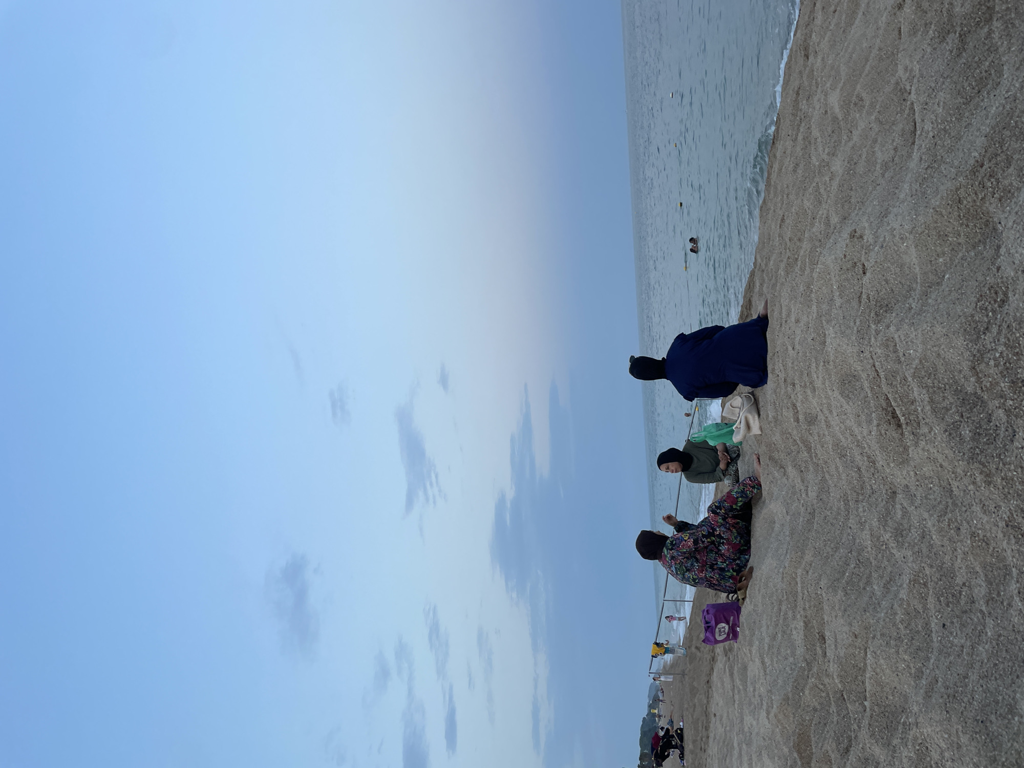 A group of three individuals, two women seated and one person facing the ocean, are on a sandy beach in Canet de Mar, Spain. The two seated women are facing each other and appear to be engaged in conversation, with a purple bag, a green bag, and a white tote bag near them. The person facing the ocean is seated closer to the water. The sea is calm with small waves, and there are a few people swimming in the distance. The sky is a soft blue, transitioning to a lighter hue towards the horizon, with scattered clouds. The beach appears to be sparsely populated with other individuals and beach gear visible further down the shore. The overall scene depicts a tranquil evening at the beach.