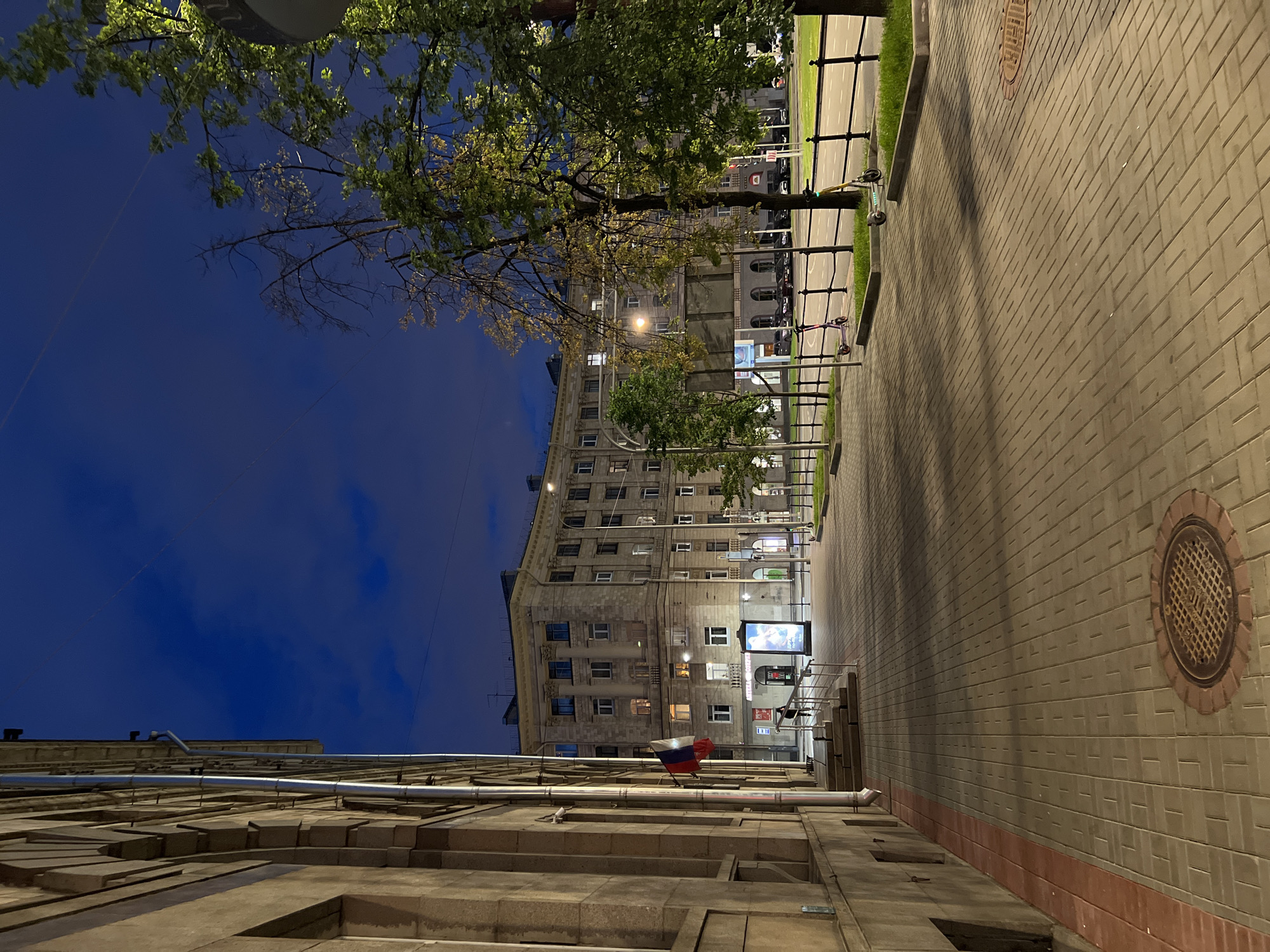 The image depicts a nighttime street scene in an urban environment. On the left, a light-colored, multi-story building with a textured facade features arched windows and a downspout. A Russian flag hangs from a pole near the entrance. A paved walkway with a red border runs alongside the building. A manhole cover is visible on the walkway.
The street itself is paved with interlocking bricks. Across the street, a large, light-colored building with classical architectural elements, including columns, rises several stories. This building has numerous windows, some of which are illuminated. Streetlights illuminate the scene.
To the right of the street, a sidewalk area is separated from the road by a black metal fence and a strip of grass. Several trees, some with green leaves and others bare, are present. Parked cars are visible behind the fence. Two electric scooters, one purple and one yellow-green, are parked on the grass area near the fence. The sky is a deep blue, indicating twilight or night. The overall impression is a quiet, well-lit urban street at dusk or night.