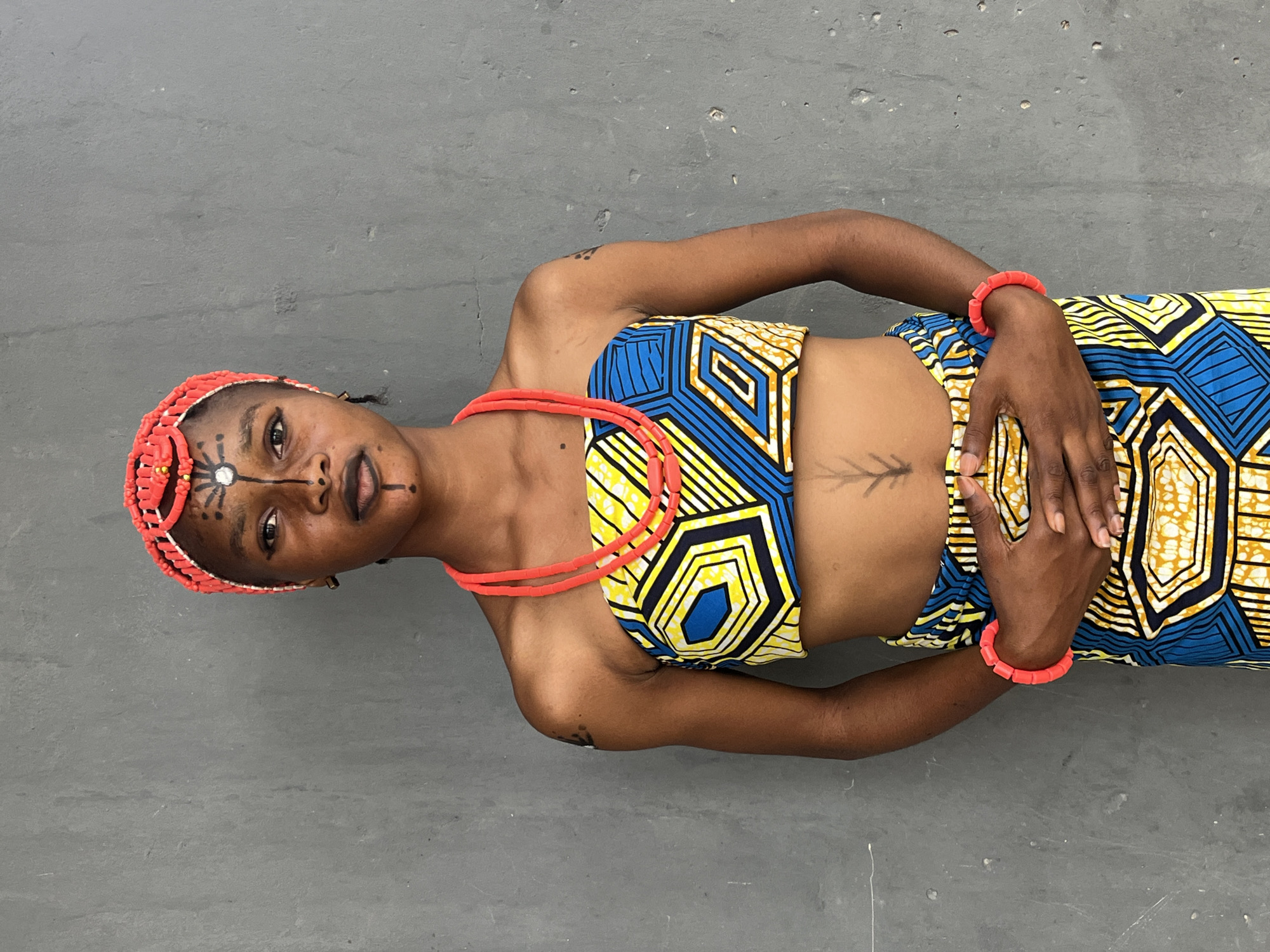 A person lies on a gray concrete surface. They are wearing a two-piece garment with a yellow, blue, and black geometric pattern, along with a red beaded necklace and bracelets. Their head is adorned with a red beaded headpiece, and they have face paint consisting of lines and a white circle. A tattoo is visible on their chest. The location is unknown.