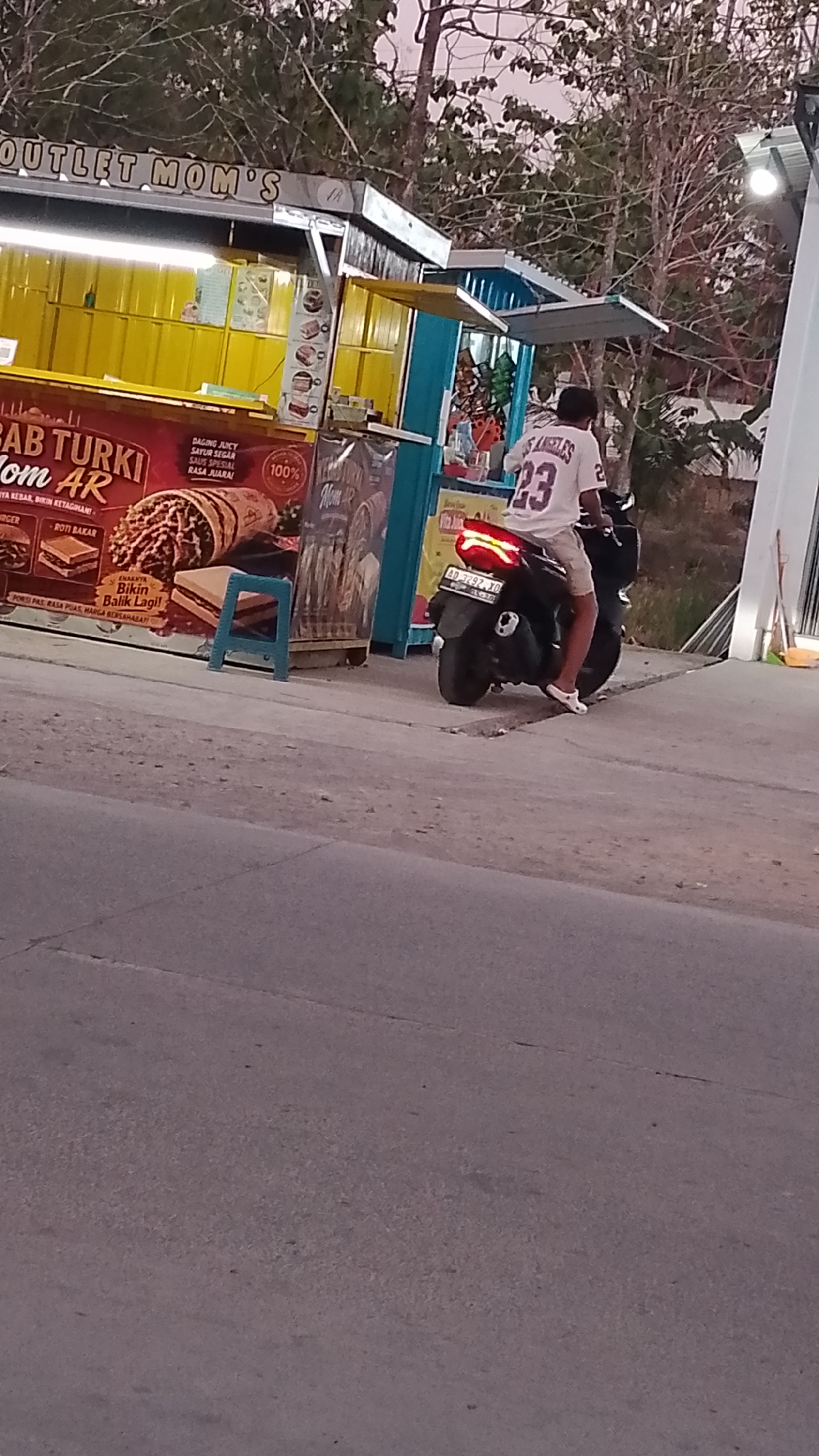 In Karanglangu, Indonesia, an individual is seated on a motorcycle with their back to the camera. They are wearing a white t-shirt with "LOS ANGELES" and the number "23" printed on it, along with tan shorts and white slip-on shoes. The motorcycle, a dark color, has a license plate reading "AD 3292 XQ". The person is positioned in front of a food stall, identifiable by a sign that reads "OUTLET MOM'S" at the top. The stall features a bright yellow facade with various advertisements, including one for "BAB TURKI Hom AR". To the right of the food stall, a tall, white structure with vertical lines is visible, and beyond that, trees and a dusky sky suggest the time is either dawn or dusk. The ground is paved and appears to be a road or pathway.