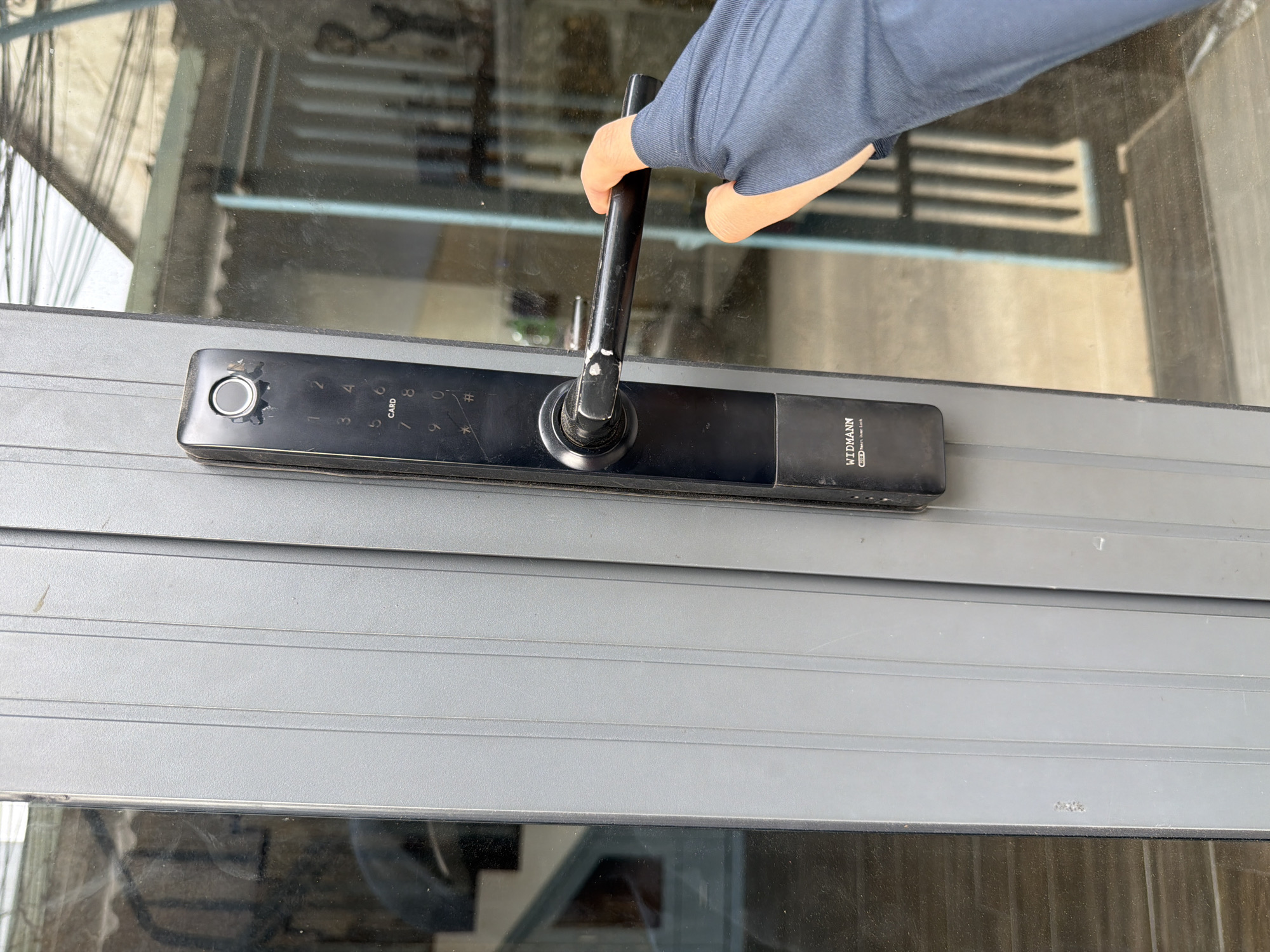 A close-up, eye-level shot captures a hand interacting with a smart door lock on a glass door or window. The scene is set in Đà Nẵng, Vietnam, under bright daylight conditions.
The main subjects are a hand, a smart lock, and the glass door/window frame. The hand, covered by a dark blue or grey long-sleeved garment, grasps the black lever-style handle of the smart lock, suggesting the person is opening or closing the door.
The smart lock is a sleek, black device mounted horizontally on a dark grey or black frame. On its left side, there's a circular fingerprint reader, which shows visible wear and some chipping around its edge. To the right of the fingerprint reader is a digital keypad with numbers 1 through 9, an asterisk (*), a hash (#), and the word "CARD." The handle itself also has visible scuff marks. On the far right of the lock, the brand name "WIDMANN" is visible, along with the text "RED Smart Door Lock."
The glass pane, which appears somewhat smudged or dirty, reflects and distorts the outdoor surroundings. Through the glass, blurred elements of an urban environment are discernible, including what look like power lines or cables and indistinct architectural features of buildings, such as horizontal slats or windows. The overall impression is one of a functional interaction with technology in an everyday urban setting.