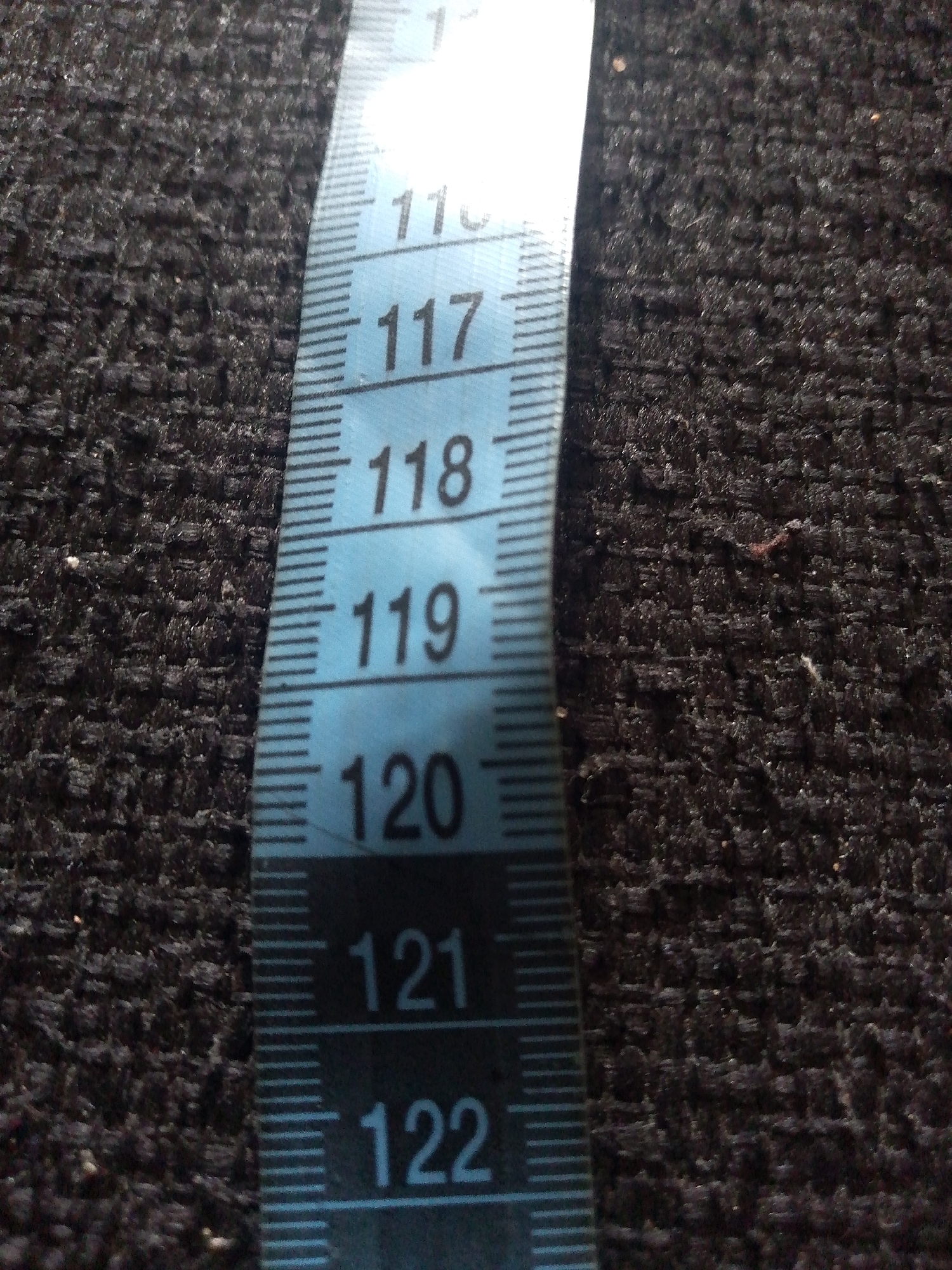 The image shows a close-up, slightly angled view of a blue and white measuring tape positioned on a dark, textured fabric. The measuring tape displays numerical markings, with the numbers 117, 118, 119, 120, 121, and 122 clearly visible. Each number is preceded by a longer horizontal line and followed by a series of shorter horizontal lines, indicating increments of measurement. The fabric appears to be a dark, woven material with a slightly raised, bumpy texture. The lighting casts subtle shadows, highlighting the texture of the fabric and the edges of the measuring tape. There are no people or any specific identifying features of a city or location visible in this close-up shot; therefore, it is impossible to determine its location in Jauro Yinu, Nigeria.