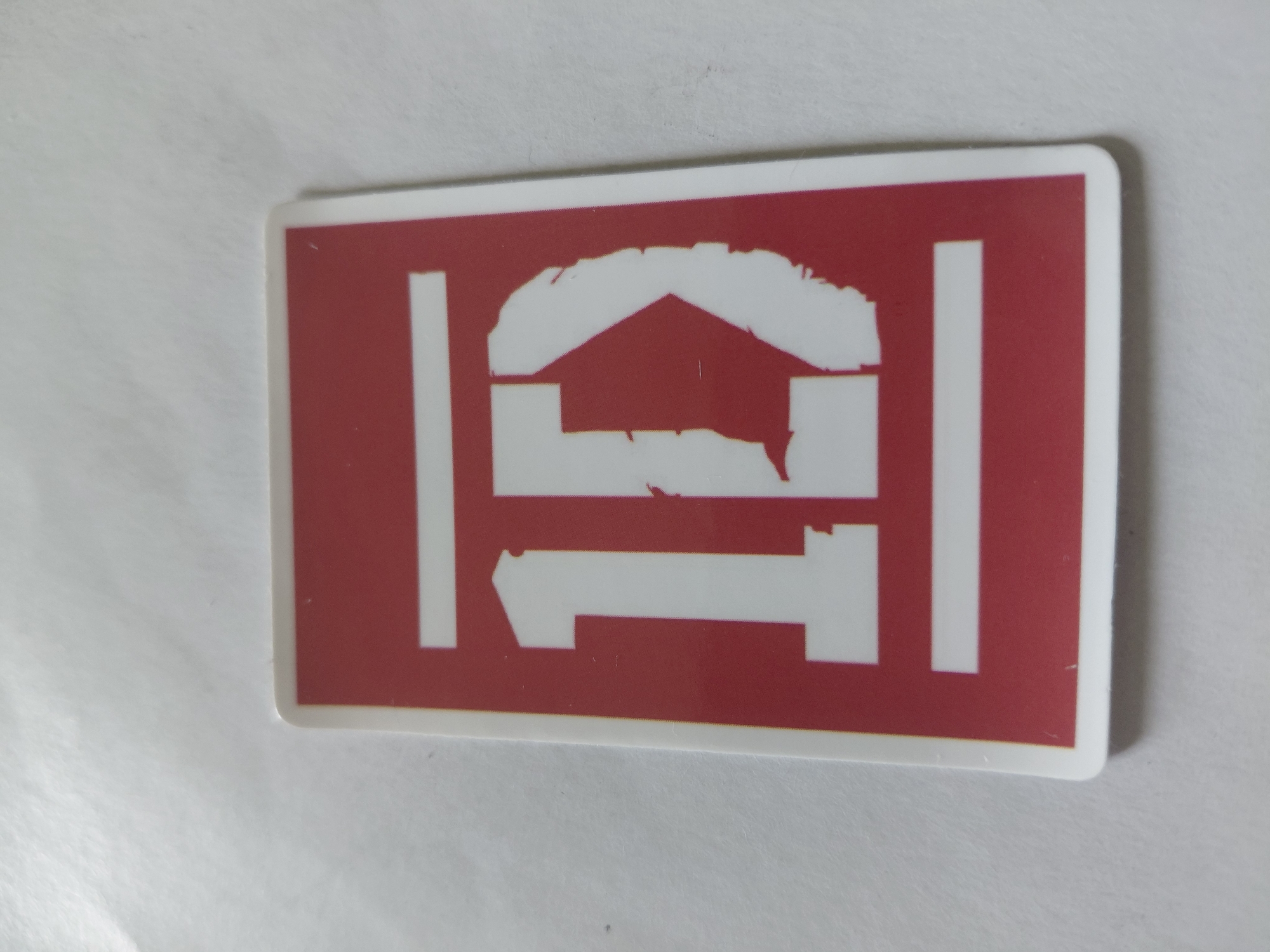 A sticker is placed on a white, textured surface. The sticker is red with a white border. It features a white design of "1D" with two horizontal white bars above and below the numbers. The surface is illuminated by overhead lighting, casting a slight shadow. There are no people or buildings visible in the image.